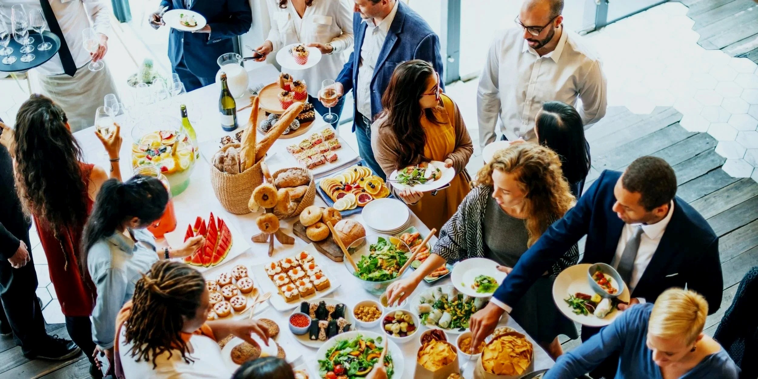 People at a buffet table filled with various foods including sandwiches, salads, snacks, bread, desserts, and drinks during a social gathering.