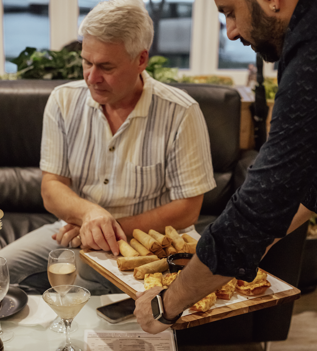 Two men serve food on a wooden platter with spring rolls, slices of quiche, and dipping sauce during a meal at a restaurant.