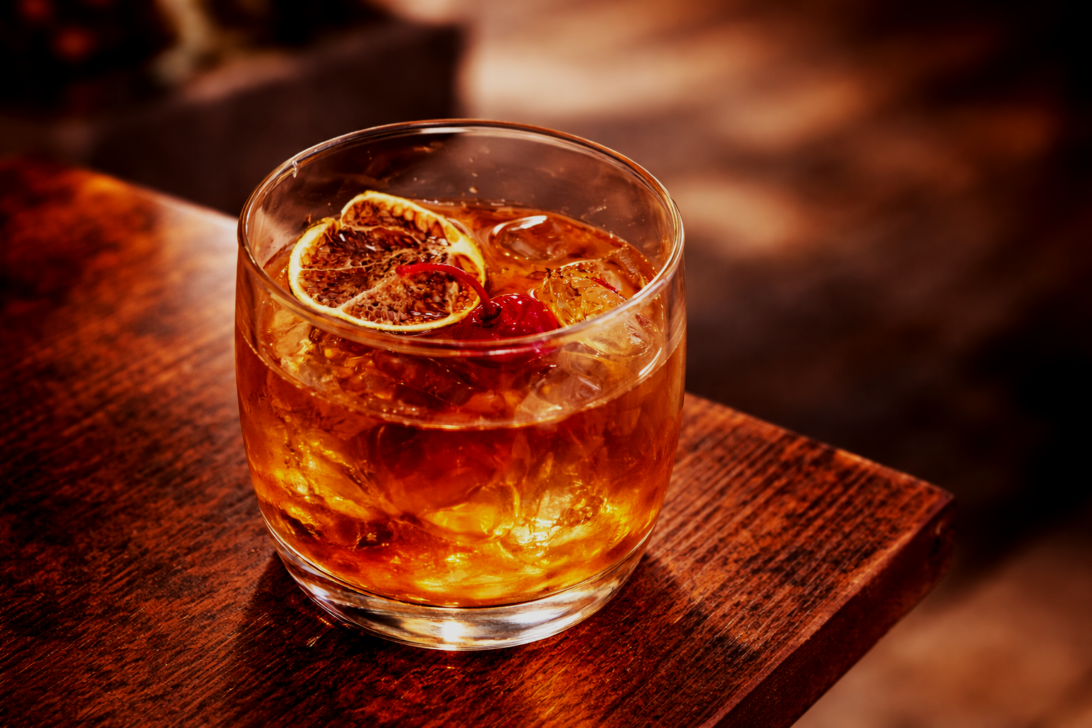 A glass of amber-colored cocktail with slices of dried orange and a cherry, served over ice, on a wooden surface.