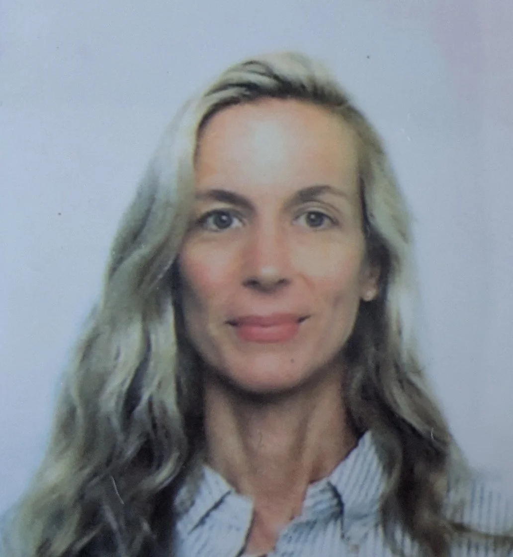 A woman with long, wavy blonde hair and blue eyes, wearing a collared shirt, standing against a plain light-colored background.