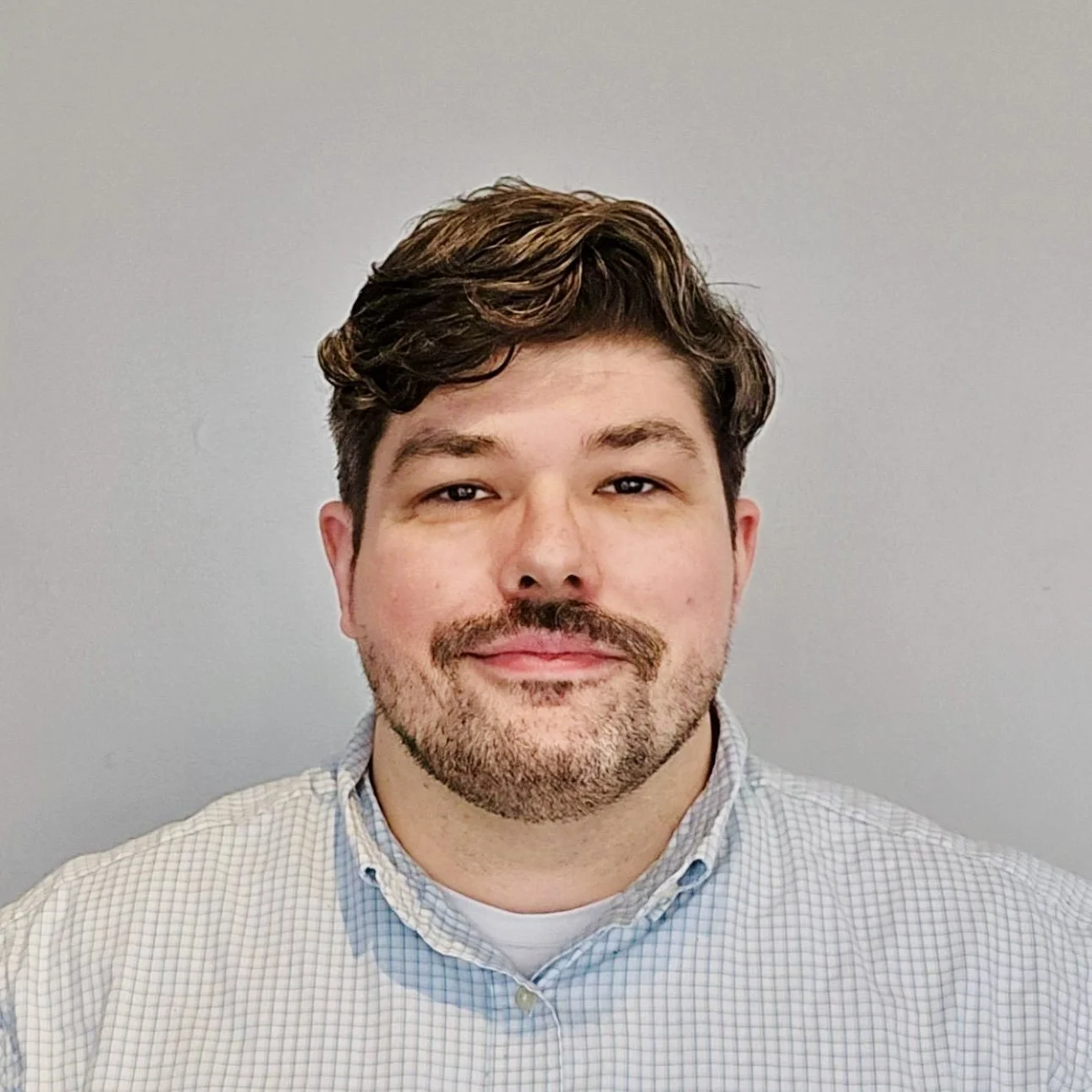 A man with glasses, a beard, and dark hair, smiling, wearing a checkered blazer, white shirt, and a patterned tie, posed against a light background.