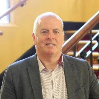 A man in a blazer posing indoors with a staircase in the background.