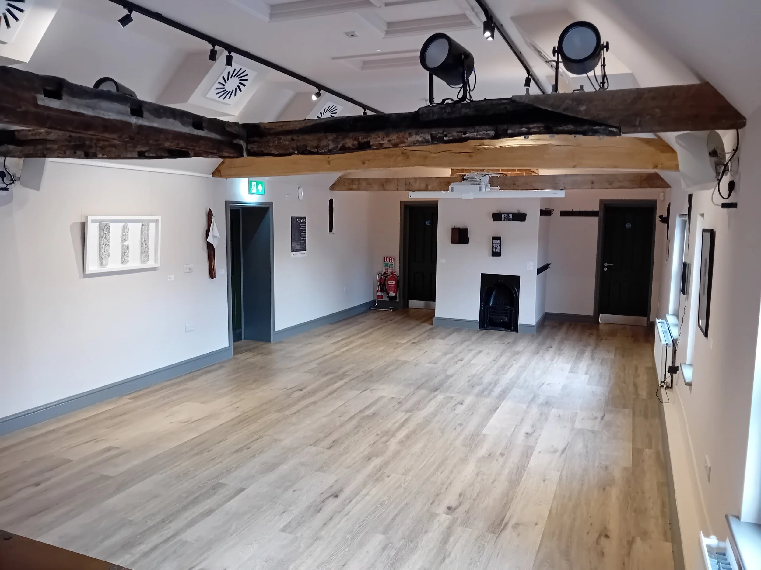 Interior of a room with light wood flooring, decorated with a white artwork on the wall and hanging wooden beams on the ceiling. Ceiling spotlights and stage lighting are also visible.