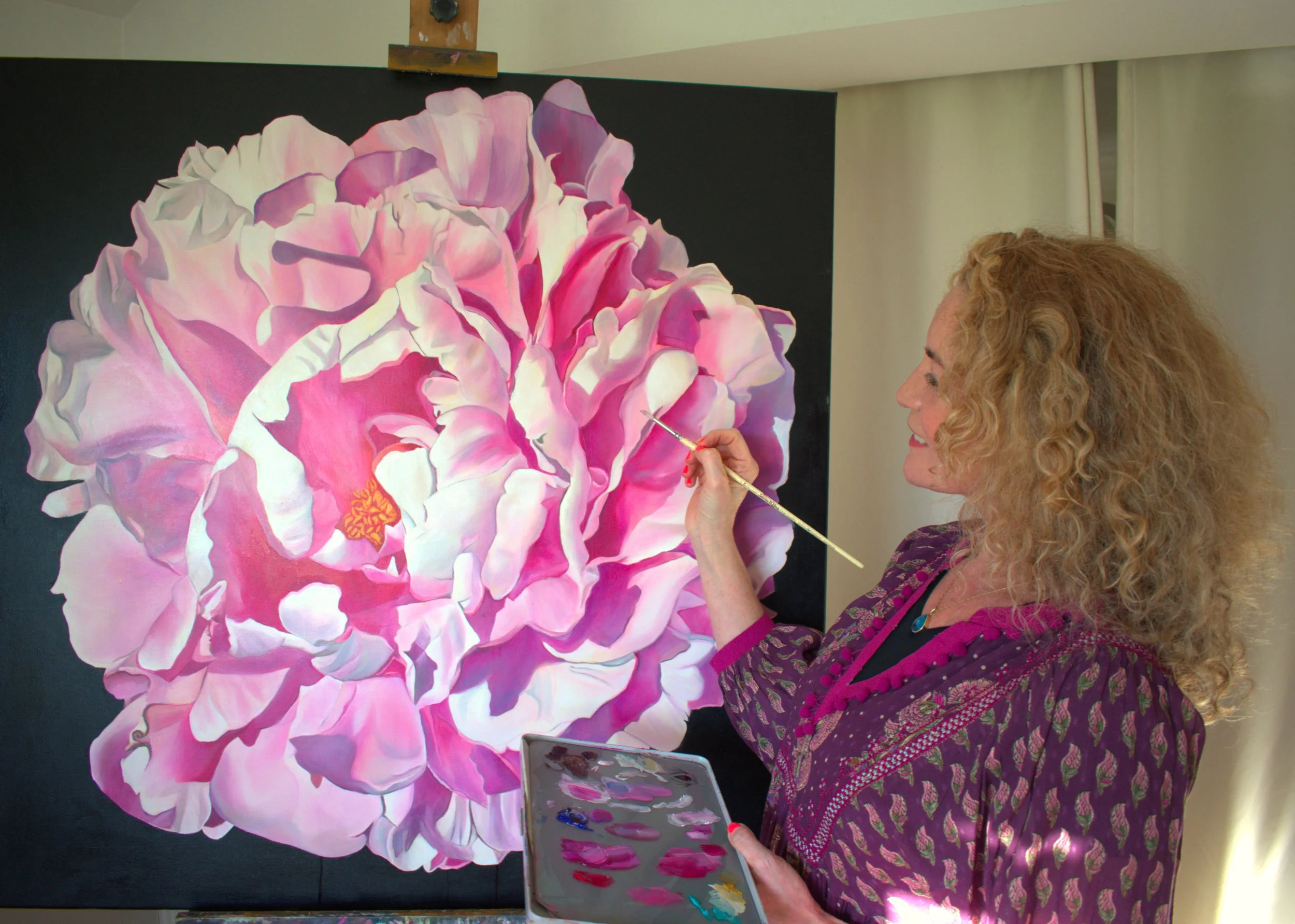 An artist painting a large pink and white flower on a black canvas. The artist, a woman with curly blonde hair, is holding a paintbrush in her right hand and a palette with various shades of pink, red, and yellow in her left hand. She is wearing a pu