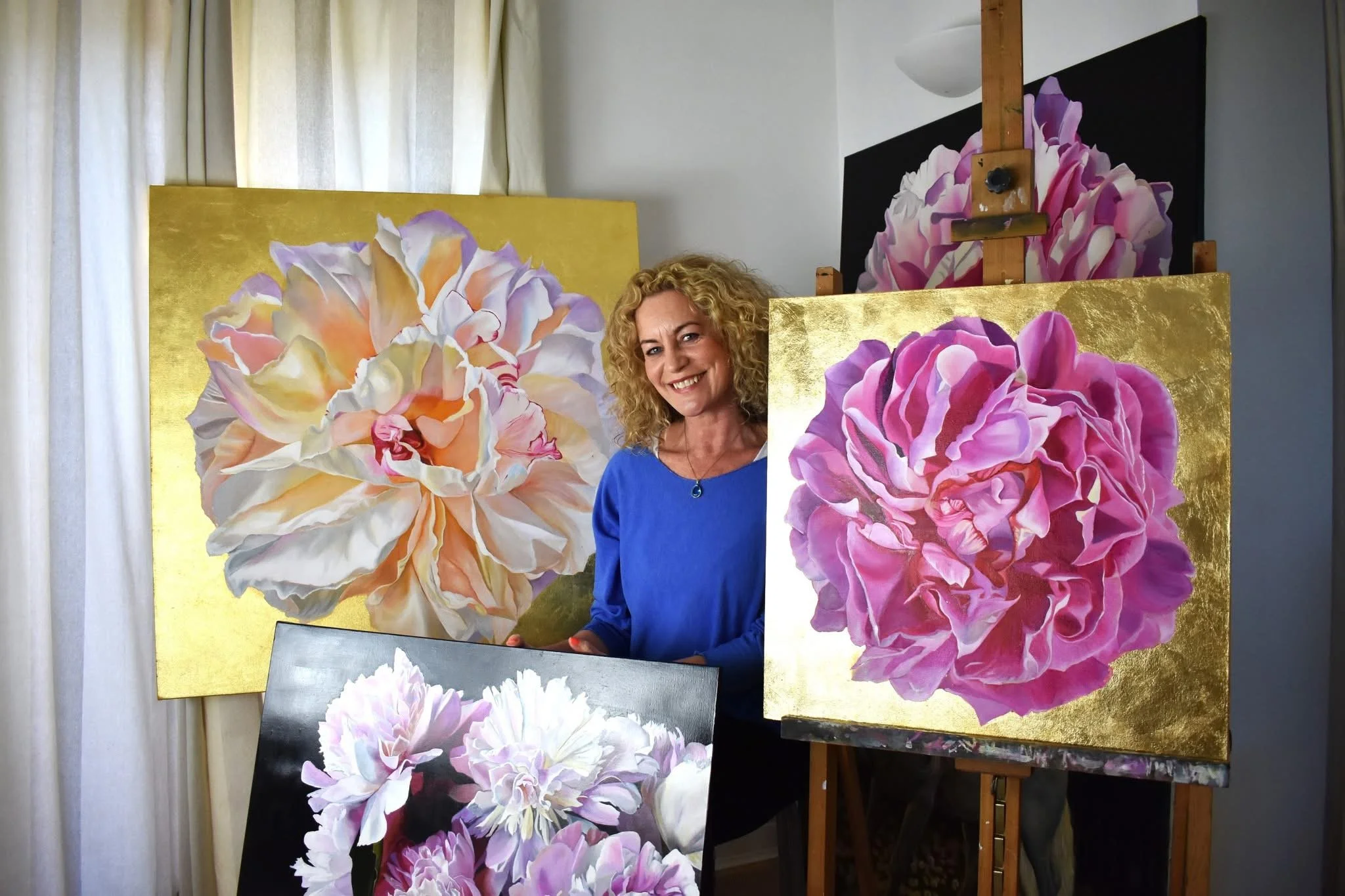 A woman with curly blonde hair wearing a blue top smiling in front of several large paintings of pink and peach flowers on gold backgrounds.