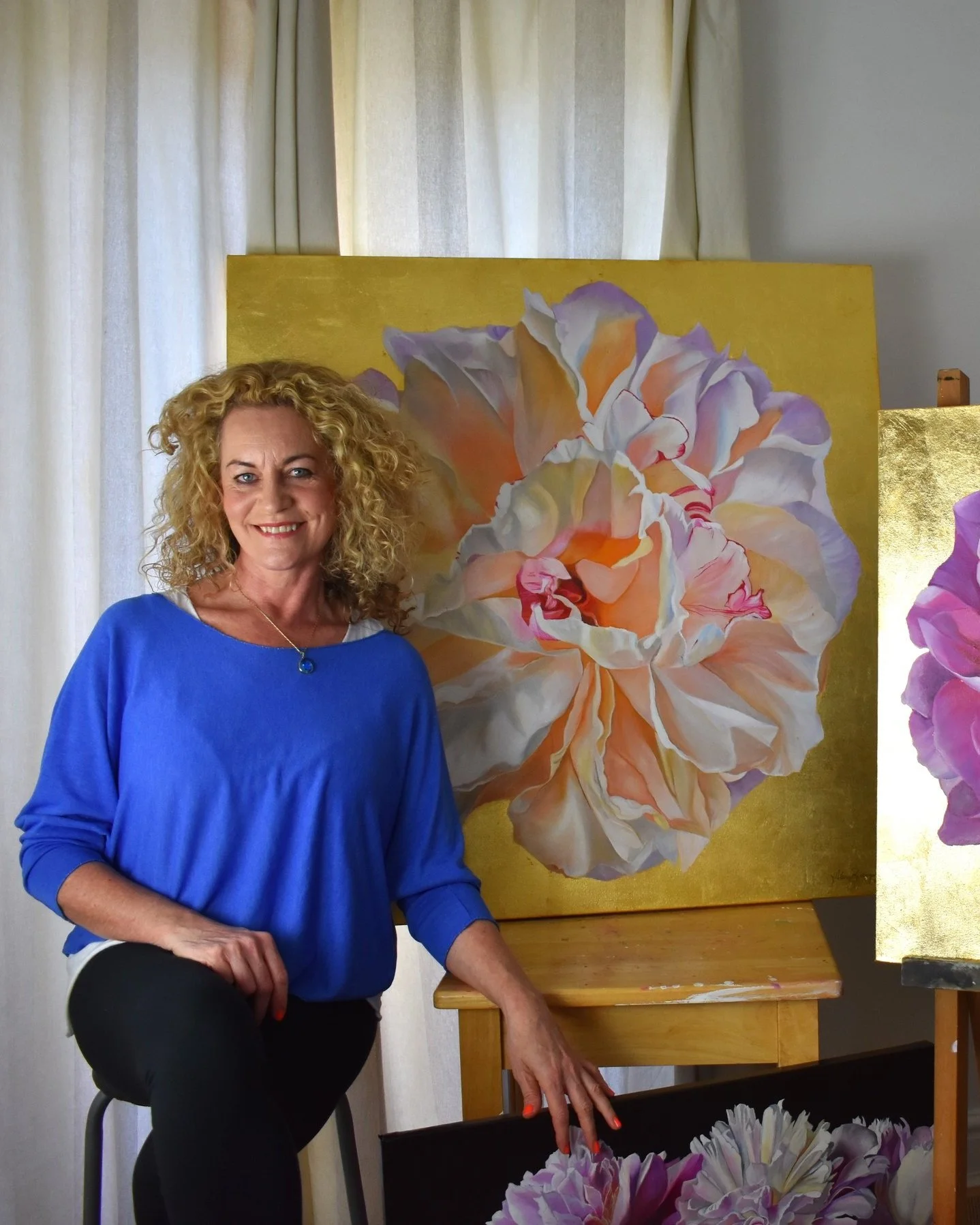 A woman with curly blonde hair smiling and sitting on a stool next to a large floral painting of a peony on a yellow background.