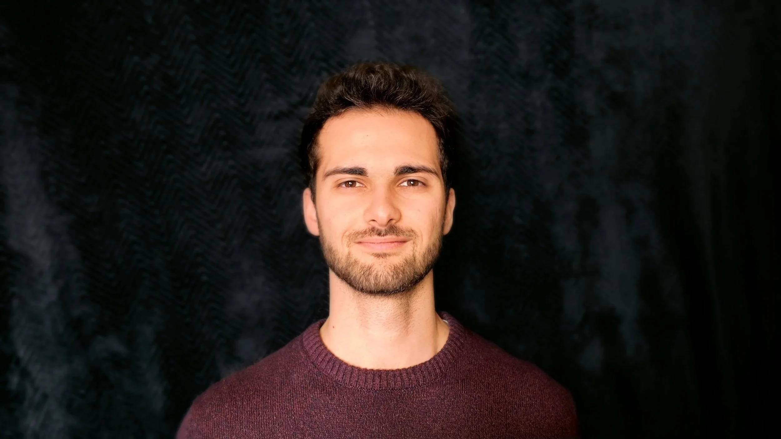 Portrait of a young man with dark hair and a beard, wearing a maroon sweater, standing against a black textured background.