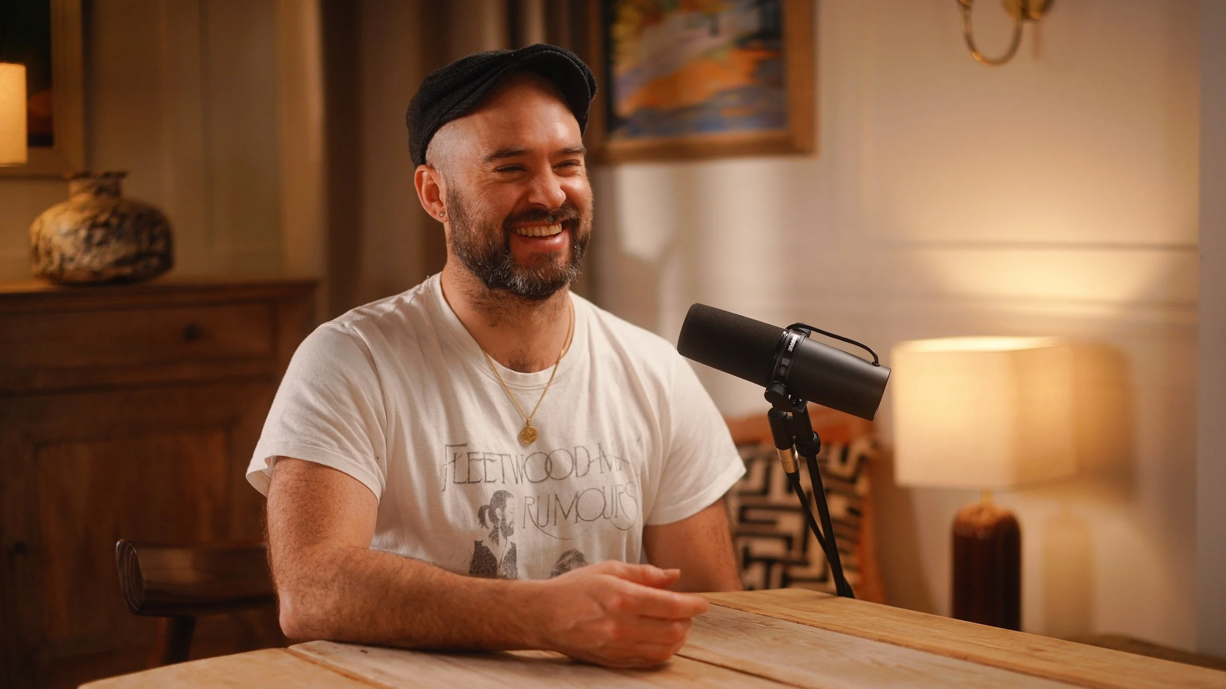 A man with a beard and mustache wearing a black cap, white Fleetwood Mac T-shirt, and a gold necklace, smiling while sitting at a wooden table with a microphone in front of him in a cozy, warmly lit room.