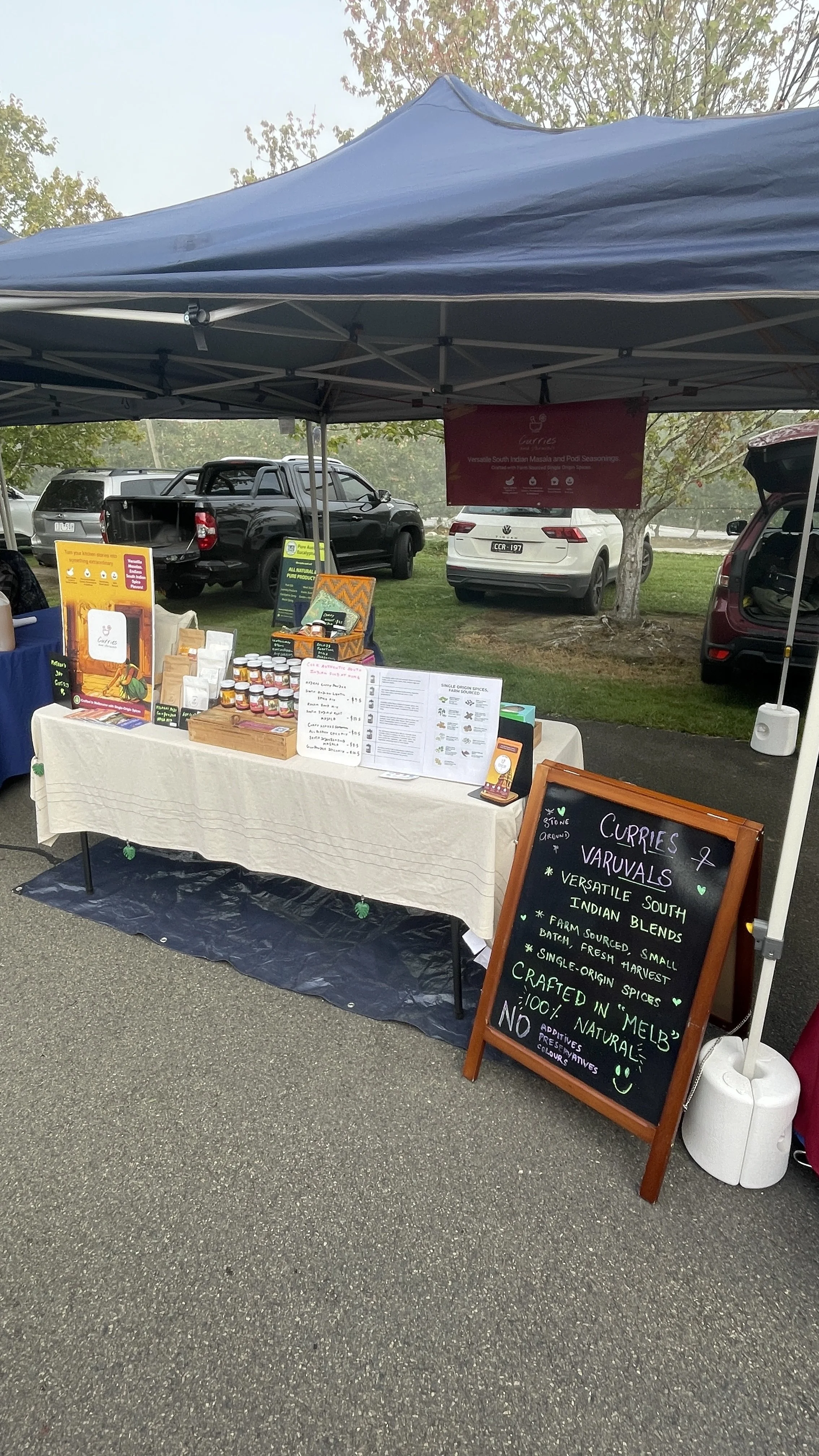 An outdoor market stall selling curries and varuvals, with a chalkboard sign highlighting the products made with farm-sourced ingredients and spices. The stall has jars of sauces or spices, displayed alongside printed menus and promotional signs, under a blue canopy with vehicles and trees in the background.