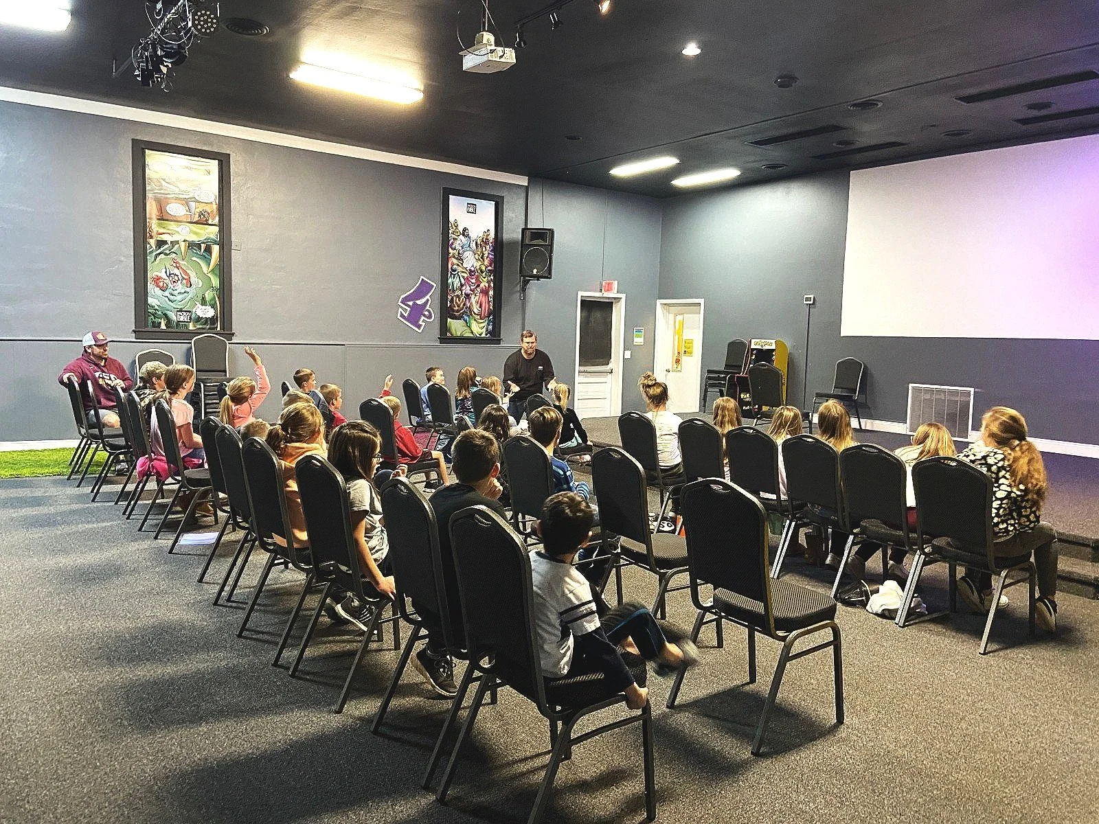 Children sitting in rows of chairs watching a presentation in a dimly lit room with a projector screen, gray walls, and colorful artwork hanging on the wall.