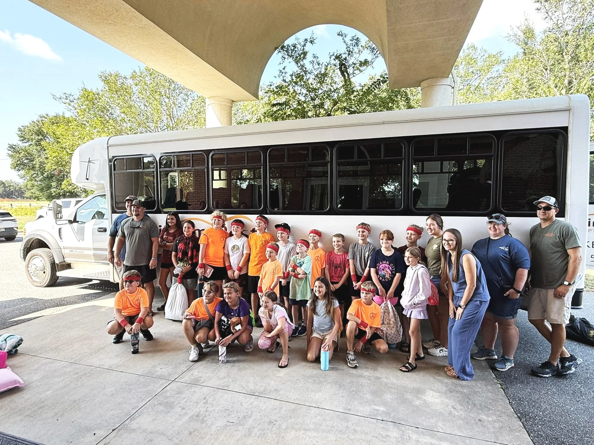 Group of children and adults standing and kneeling in front of a bus under a bridge in daytime.
