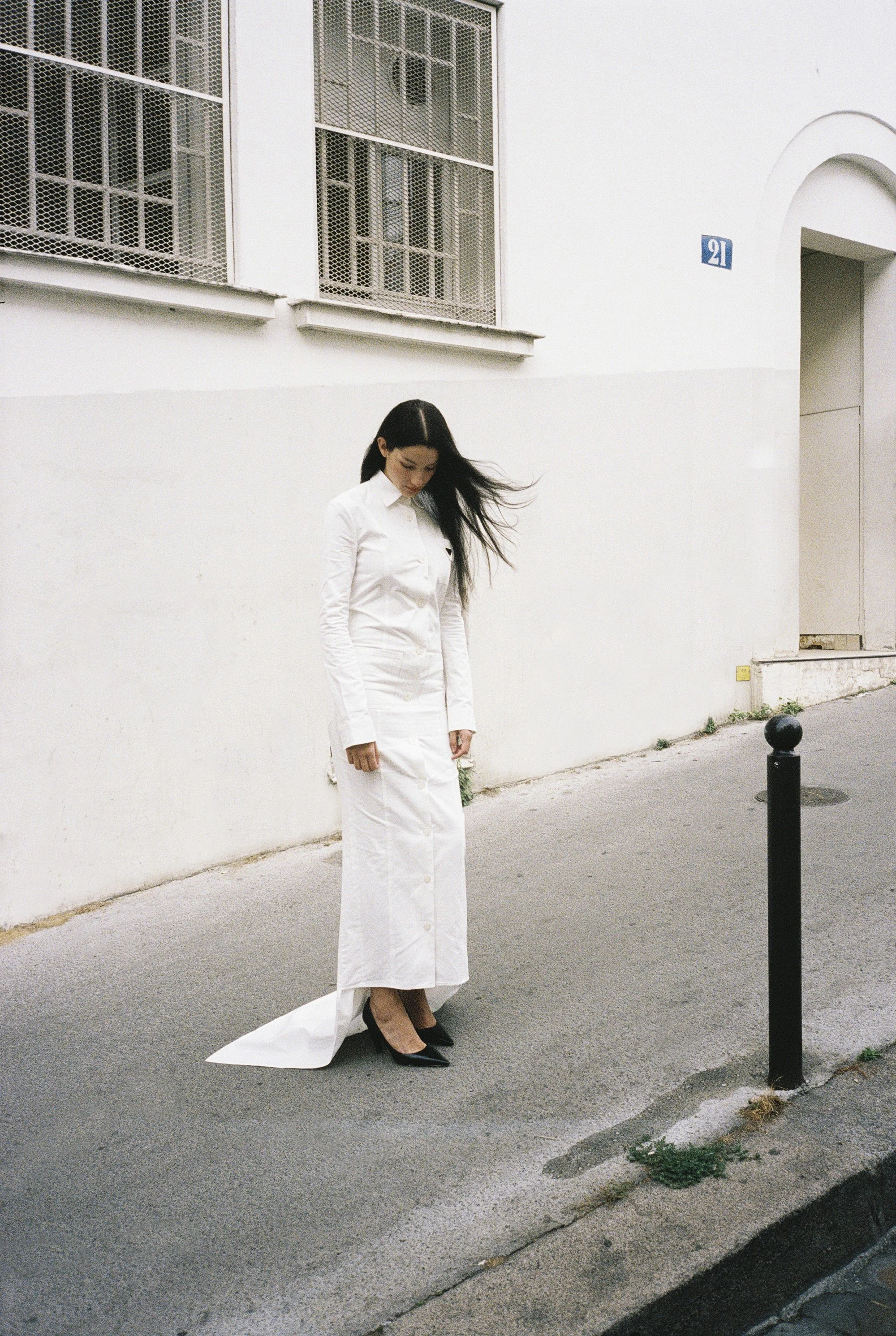 Mujer con vestido largo y zapatos negros, de cabello largo y oscuro, caminando por la calle junto a una pared blanca, en un entorno urbano.