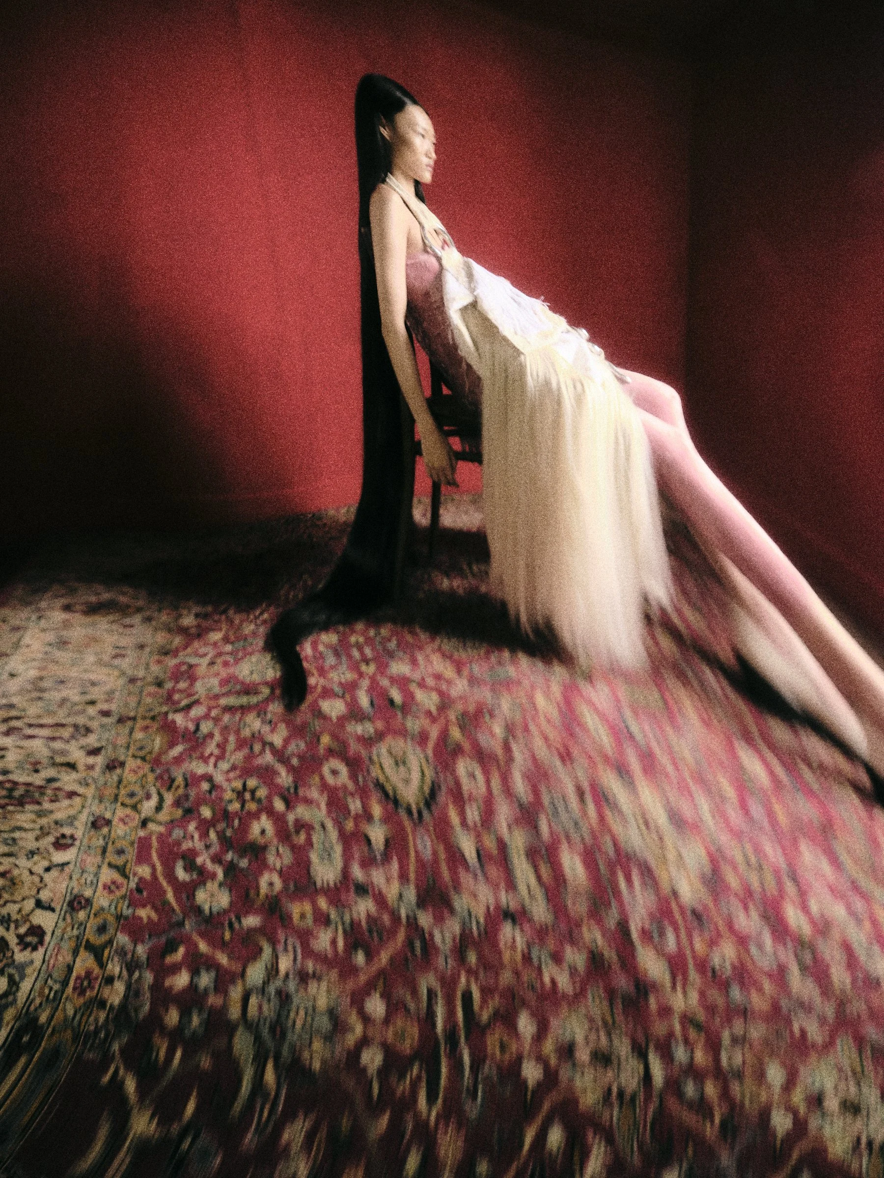 Mujer de cabello largo y oscuro, vestida con un vestido largo y fluido, sentada en una silla contra una pared roja. La imagen está ligeramente borrosa.