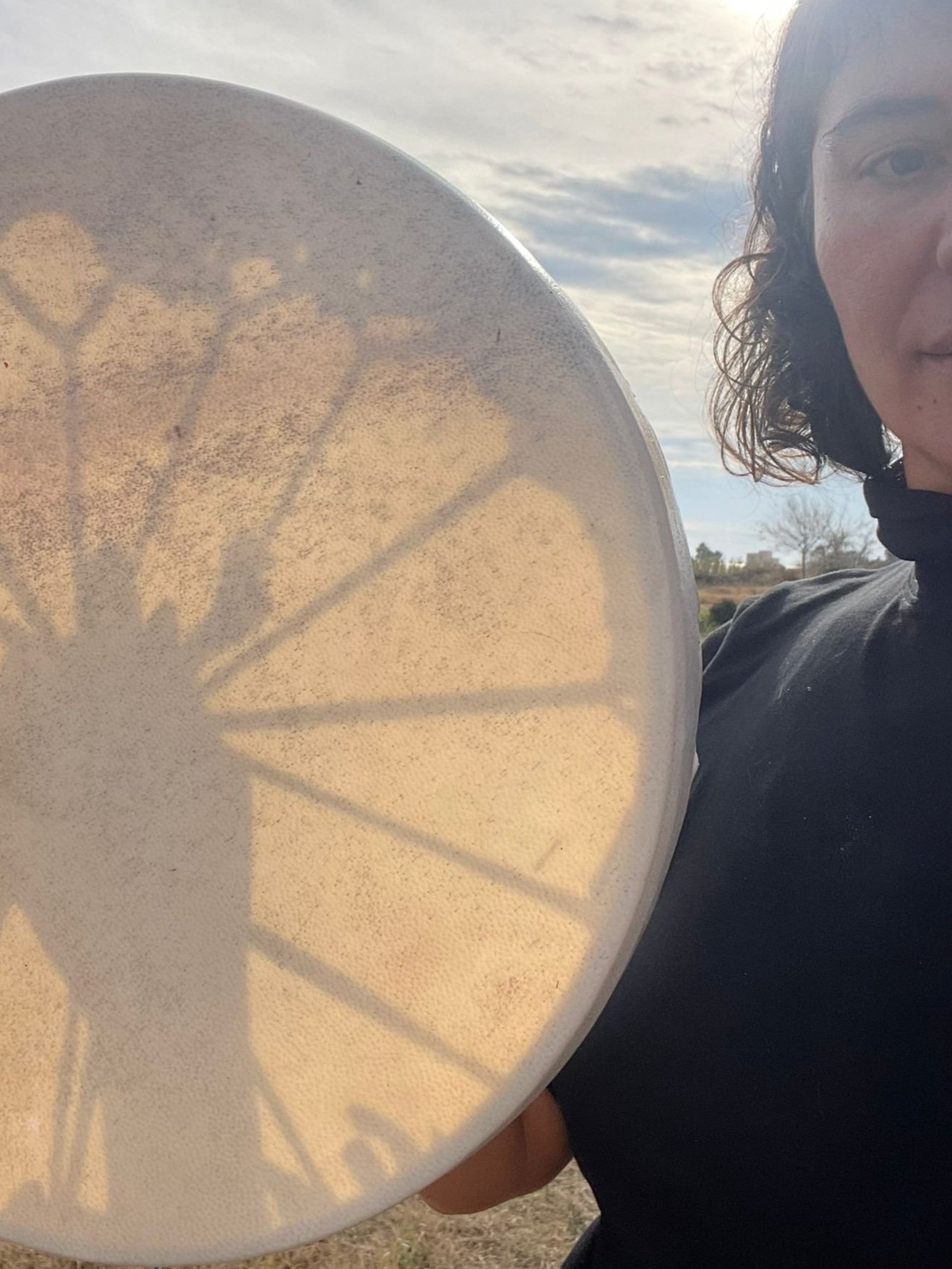 Persona sosteniendo un tambor de marco bajo el sol, proyectando sombras débilmente visibles en la superficie del tambor.