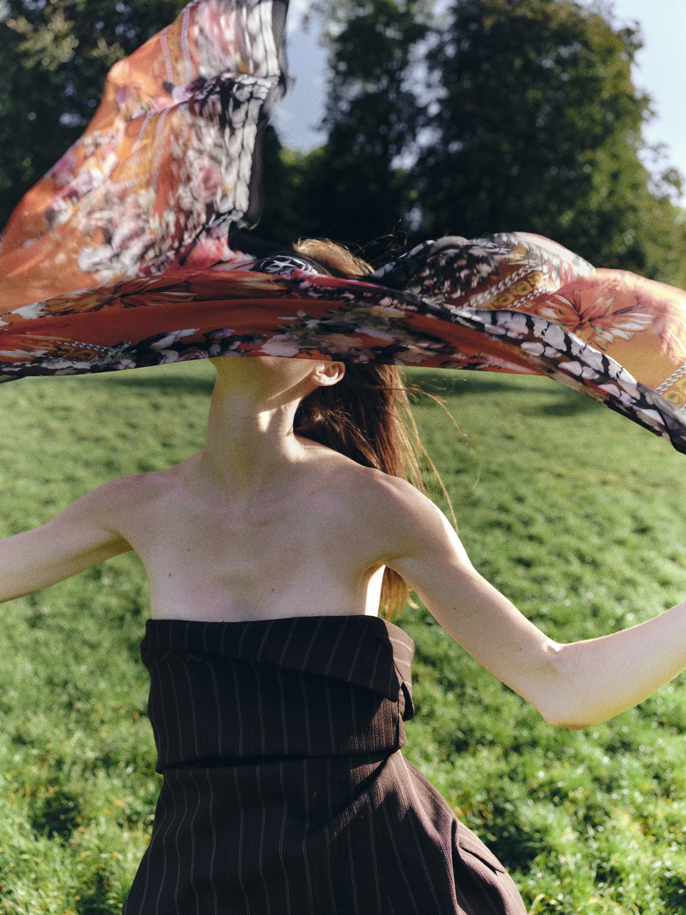 Mujer con vestido sin hombros, sosteniendo un pañuelo colorido y fluido en el aire en un parque con árboles verdes y cielo despejado.