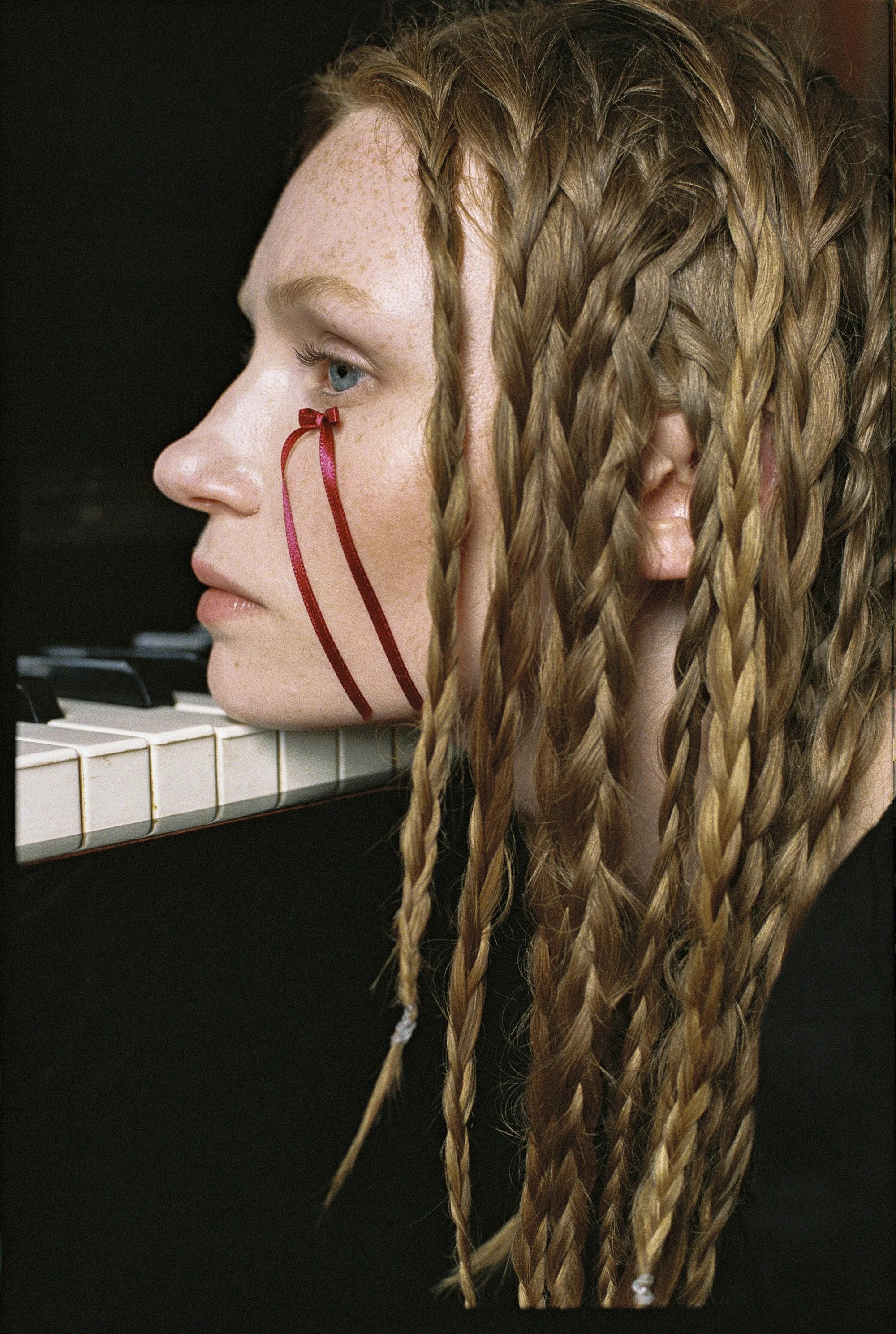 Una mujer con cabello trenzado toca un piano, con cinta roja decorativa en su rostro.