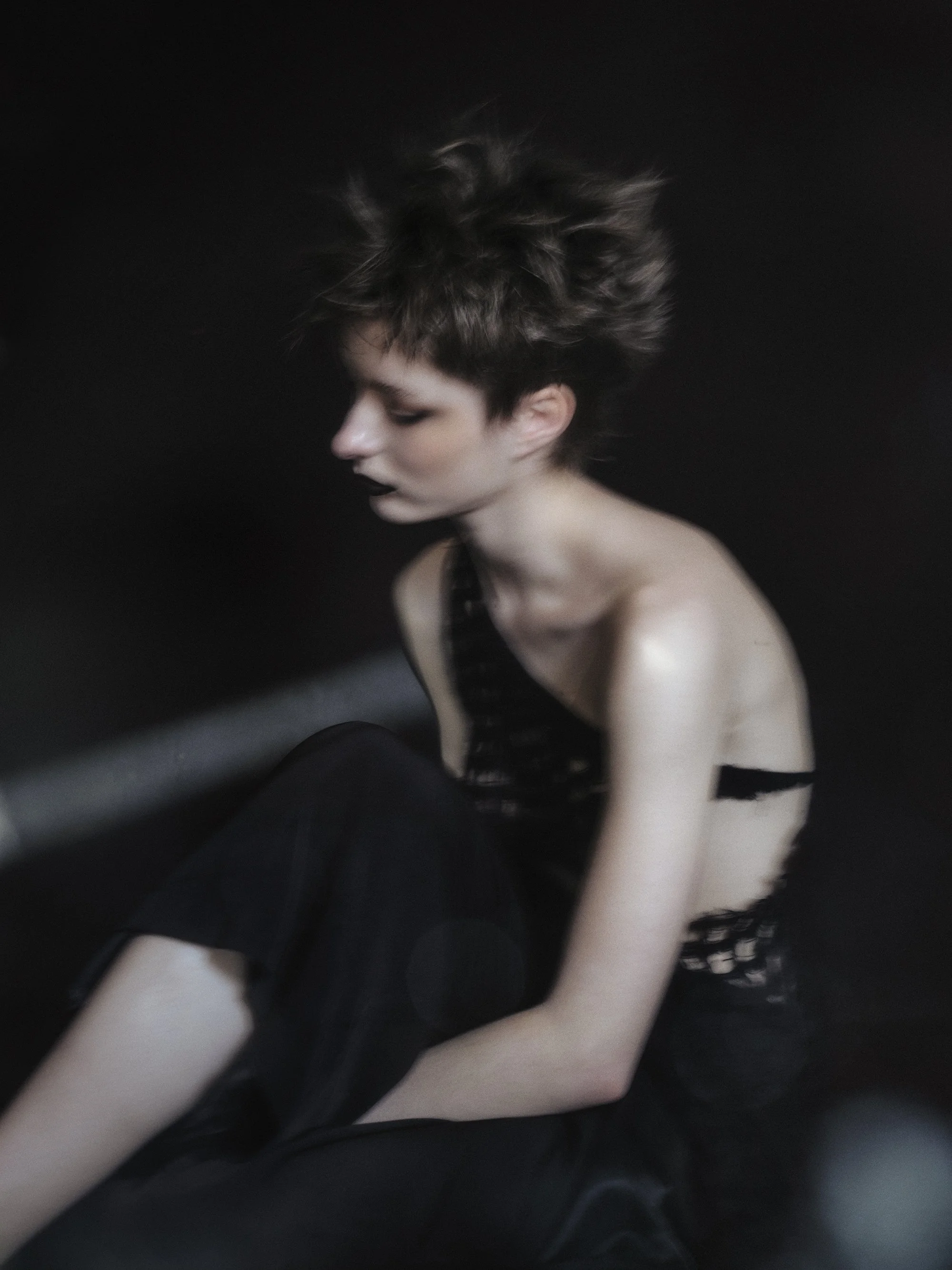 Retrato artístico de una mujer con cabello corto y desordenado, vestida con ropa negra, ubicada contra un fondo negro.