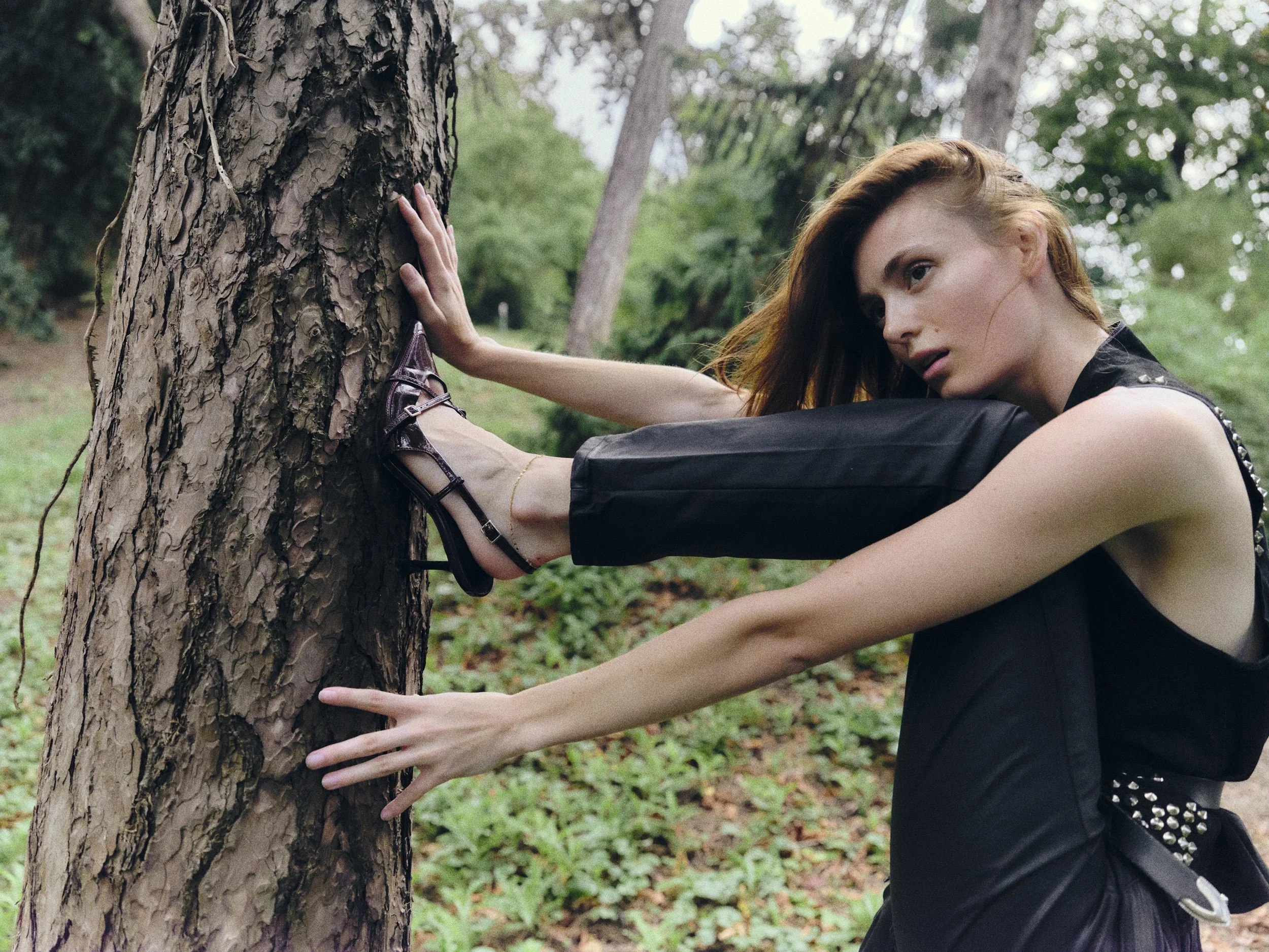 Mujer de cabello castaño claro y piel pálida, vestida con un chaleco negro y pantalones negros, está en un bosque, tocando un árbol con ambas manos, con expresión pensativa.
