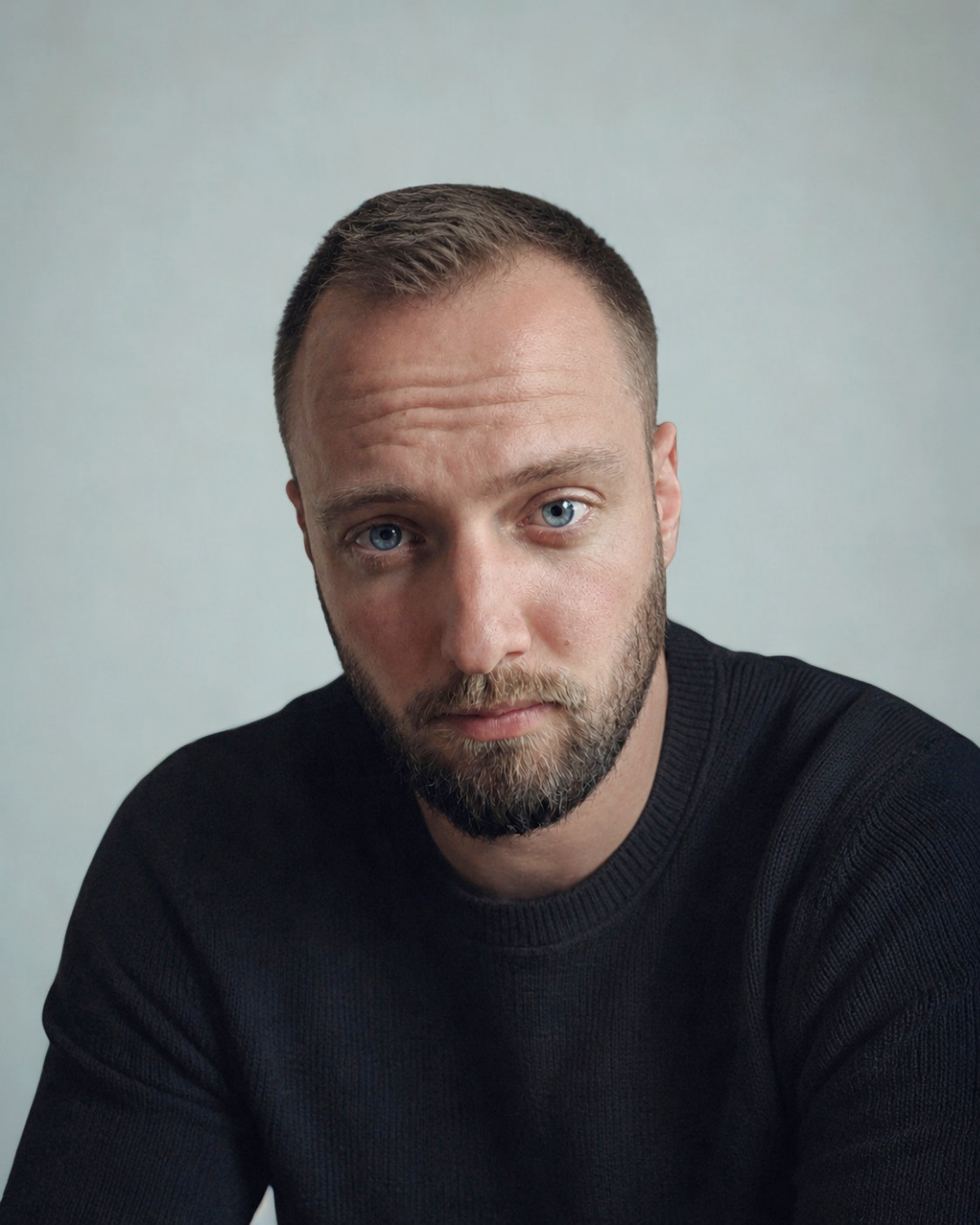 Portret van een man met blauwe ogen, kort haar, een baard en een zwart shirt, zittend voor een lichte achtergrond.