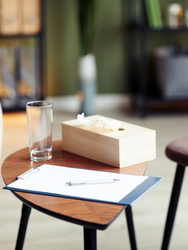 Tavolo rotondo con un blocco notes, penna, bicchiere d'acqua e una scatola di cartone con fazzoletti, ambientato in uno studio o ufficio