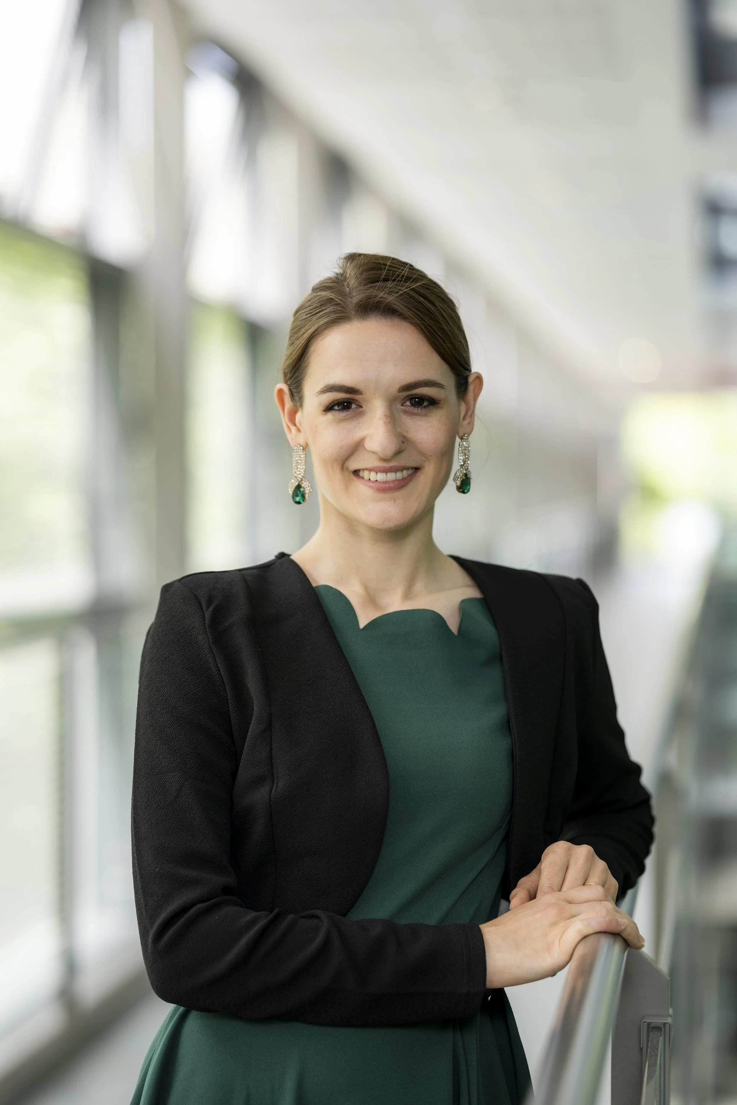 Dr Ellie Atayee-Bennett is shown smiling with brown hair, earrings, and a black blazer over a green dress, standing in a modern indoor setting with large windows.