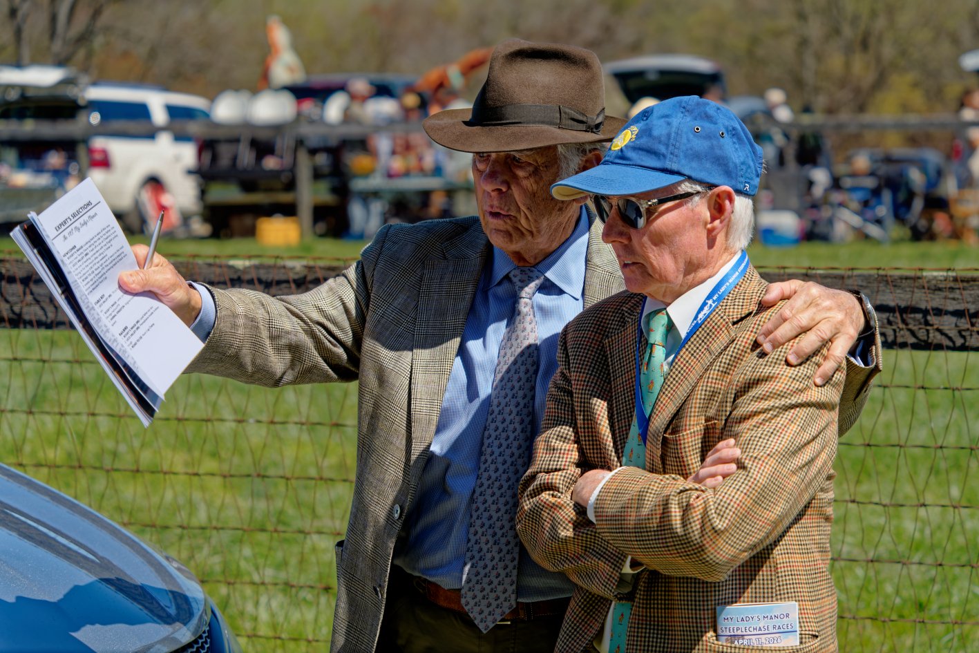 Two elderly men in plaid suits and ties, with one wearing a brown fedora hat and the other a blue baseball cap and sunglasses, standing outdoors near a fence at a racing event in the daytime. One man is holding papers and pointing, while the other ha