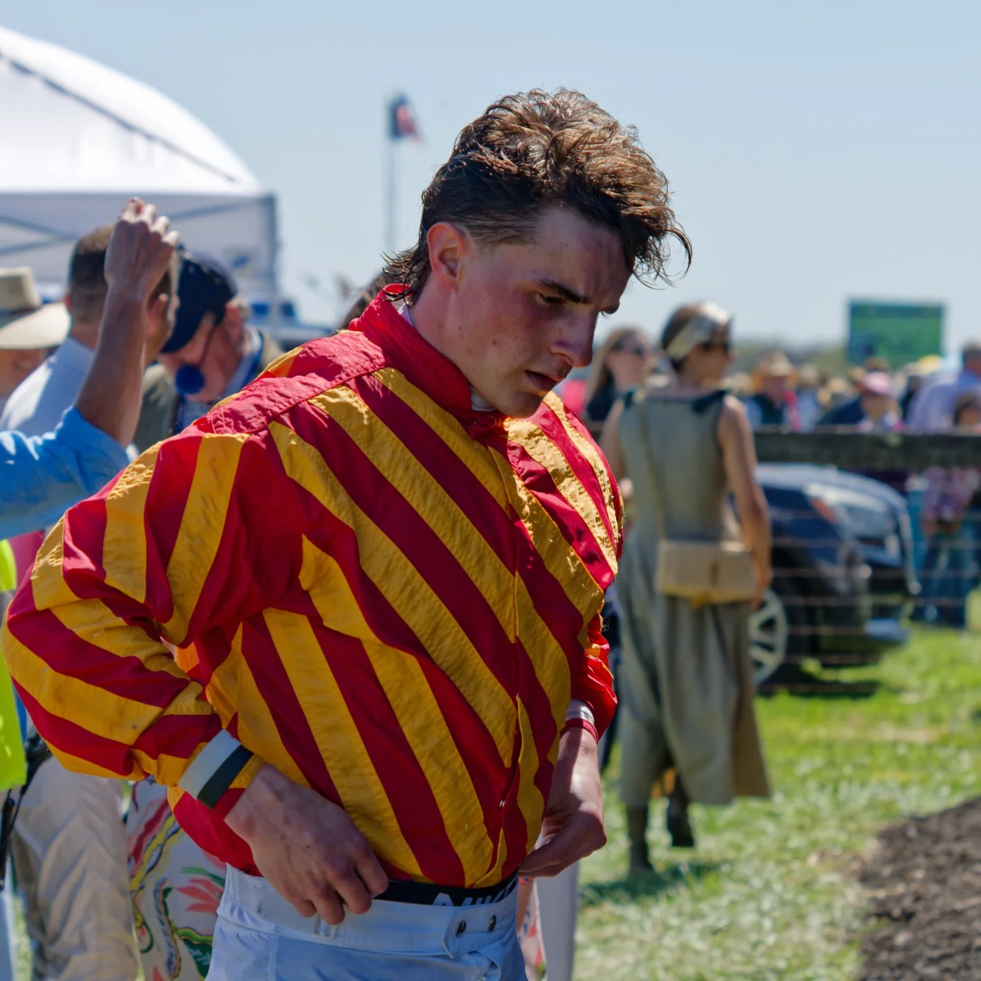 A young male race car driver in a red and yellow racing suit adjusting his belt before a race, surrounded by spectators and race officials outdoors.