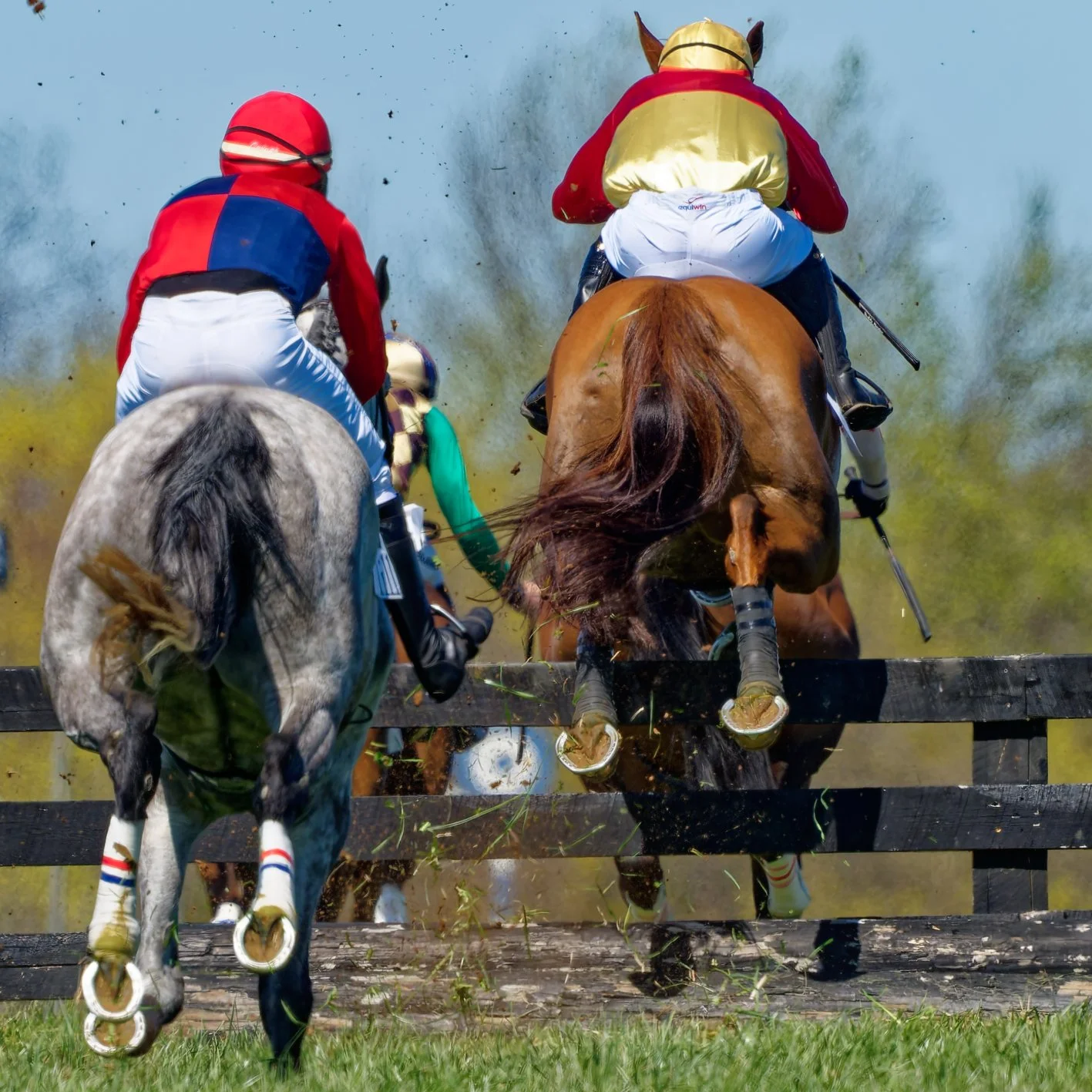 Horses and jockeys jumping over a hurdle during a horse race, with dust and grass flying in the air.