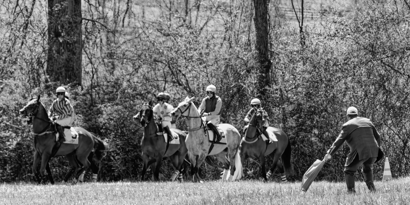 Group of five horseback riders in a line, wearing helmets, being guided by a man in a suit with a flag on a grassy field with trees in the background.