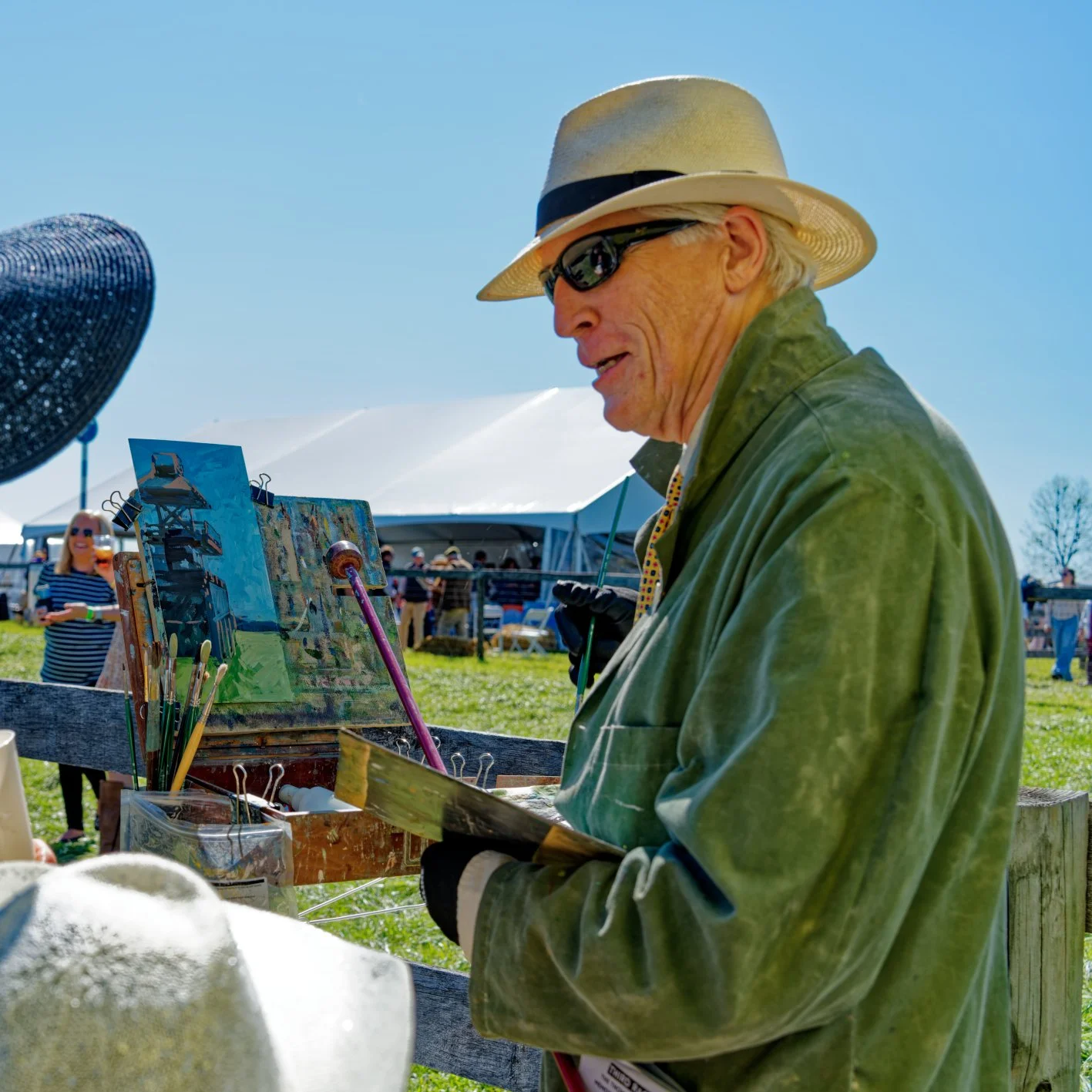An elderly man wearing a beige hat, sunglasses, and a green jacket is painting outdoors at an art fair or festival on a sunny day. Behind him, there are tents and other people, some of whom are also enjoying the event.