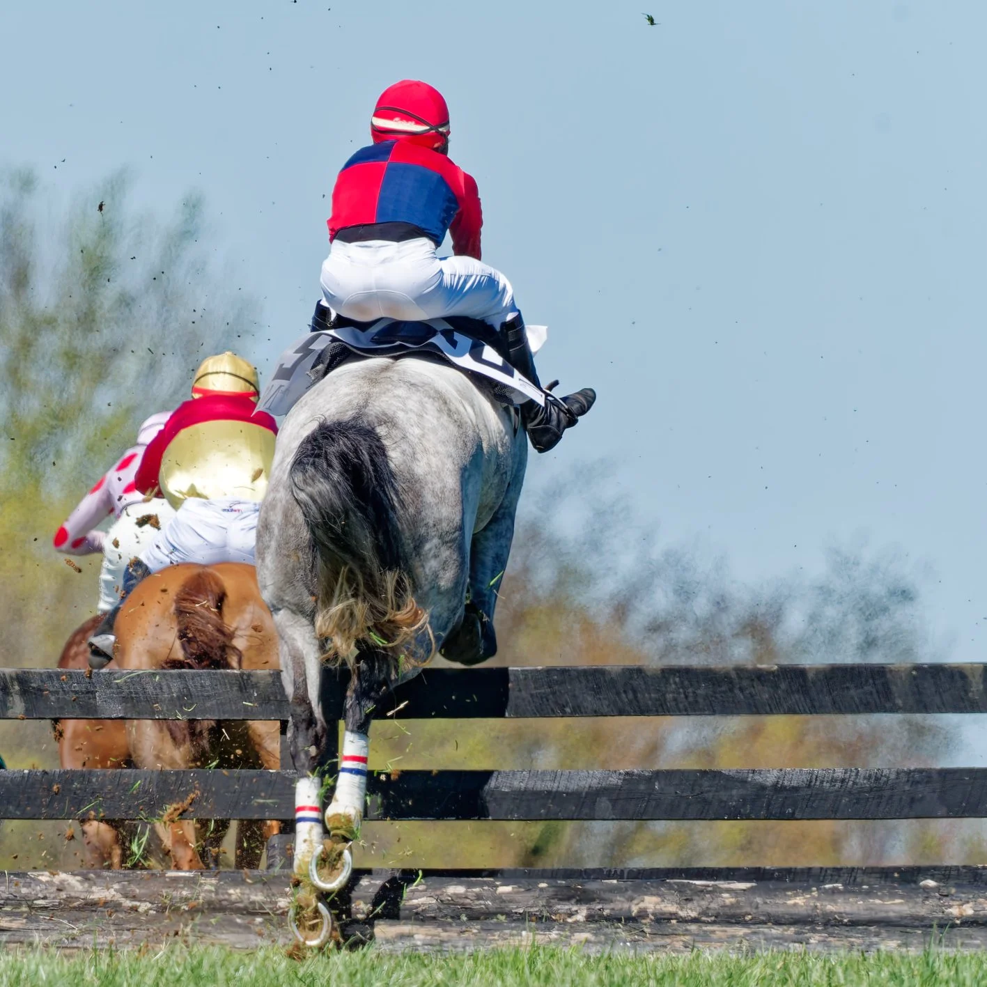 Jockeys riding horses over a wooden fence during a race, with dust and grass flying.