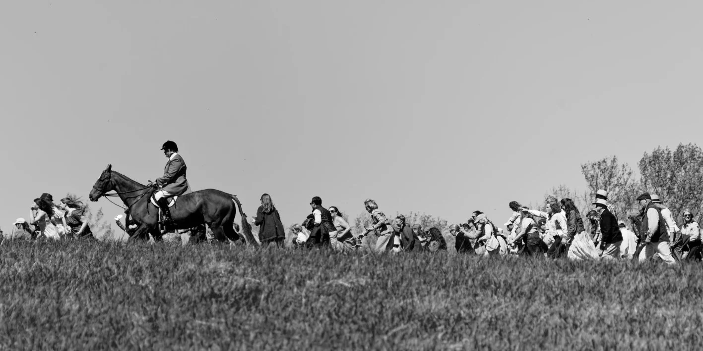A historical reenactment scene in black and white, featuring a man on horseback leading a procession of people in period clothing across a grassy field with trees in the background.