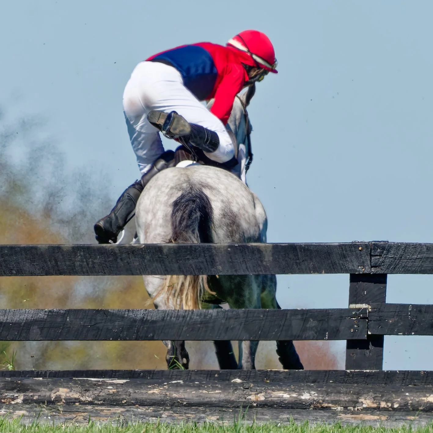 A jockey riding a grey and black horse jumping over a wooden fence during a horse race.