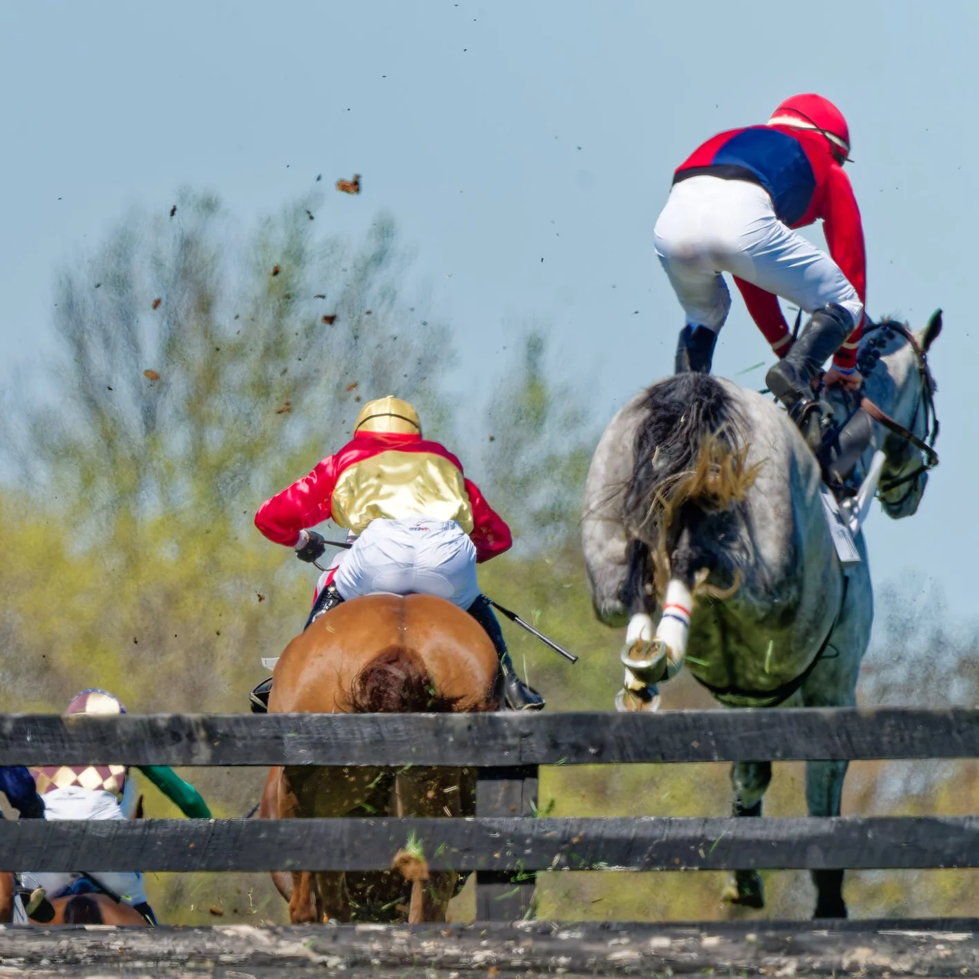 Two jockeys riding horses over a wooden fence during a horse race, with dust and debris in the air and trees in the background.