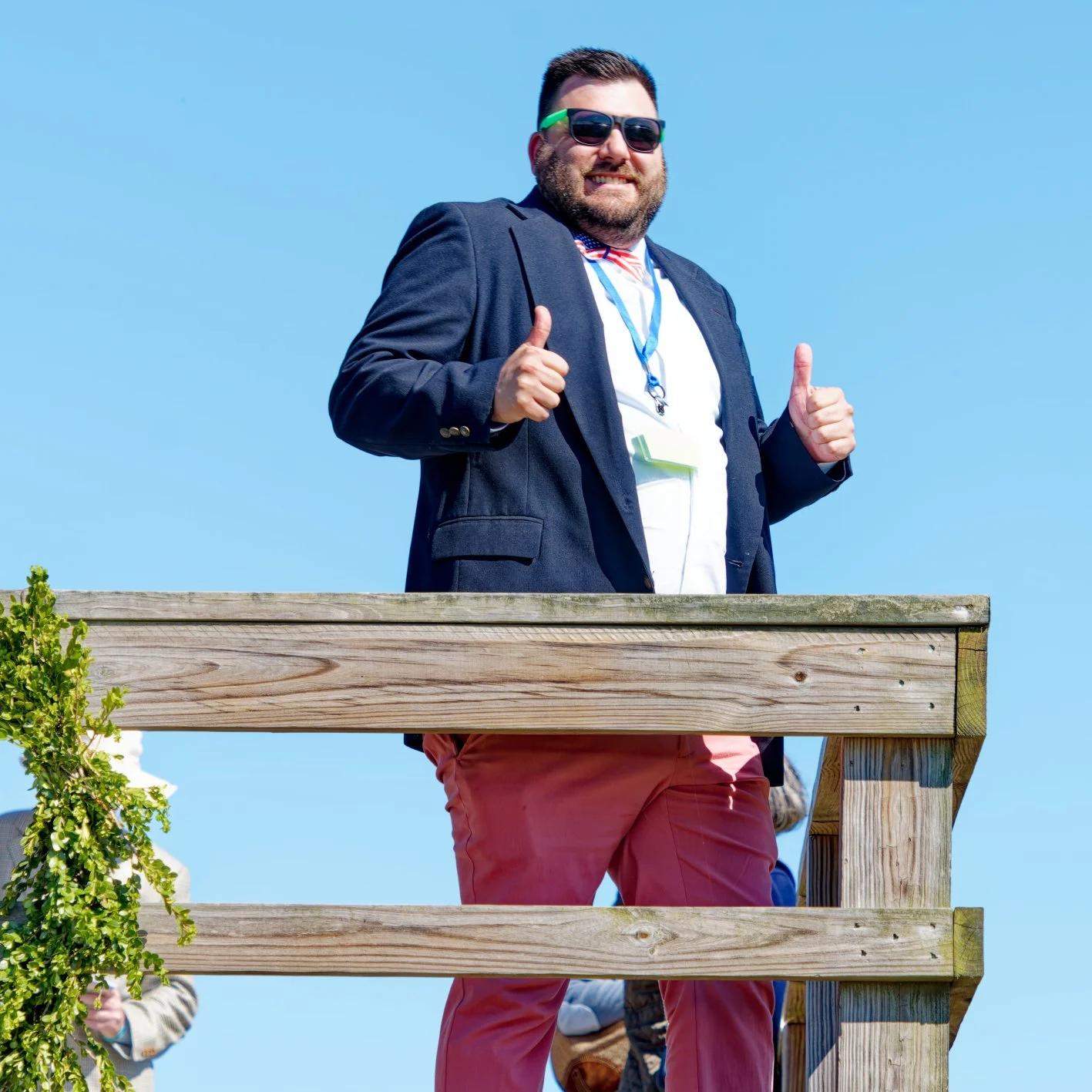 A man smiling and giving thumbs up while standing on an outdoor wooden platform, wearing sunglasses, a blazer, and red pants, with a blue sky in the background.