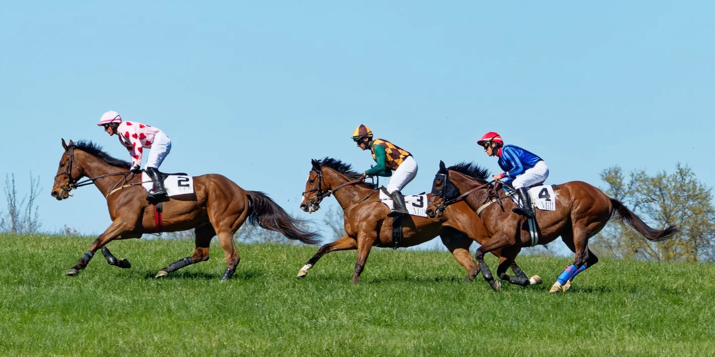 Three racehorses with jockeys in colorful uniforms competing on a grassy field under a clear blue sky.