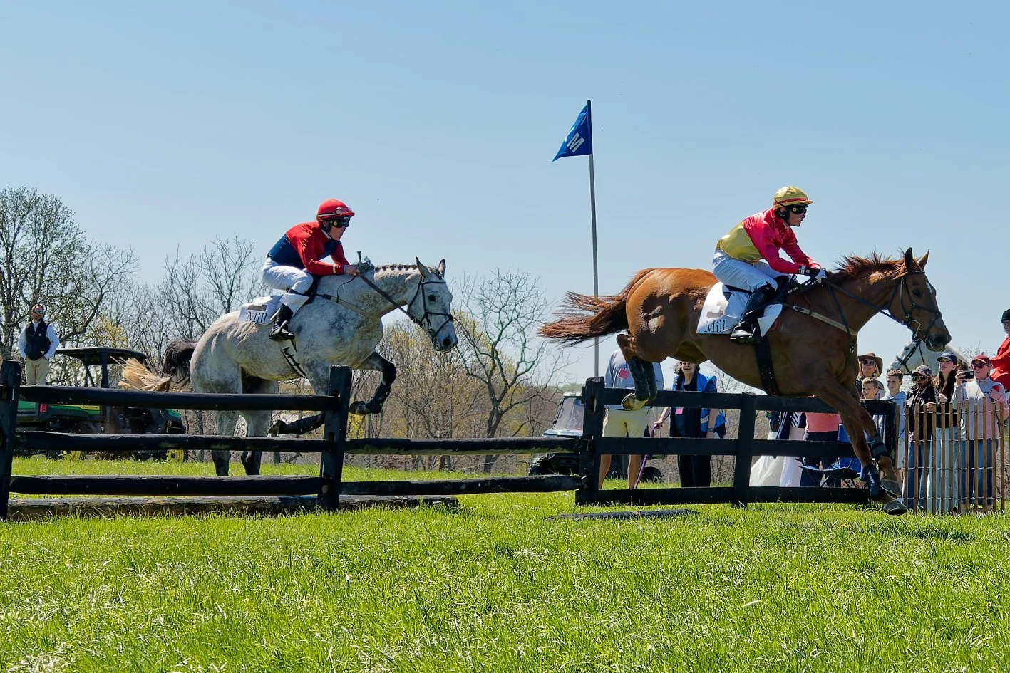 Two jockeys riding racehorses over a cross-country fence during a horse race, with spectators watching in the background on a sunny day.