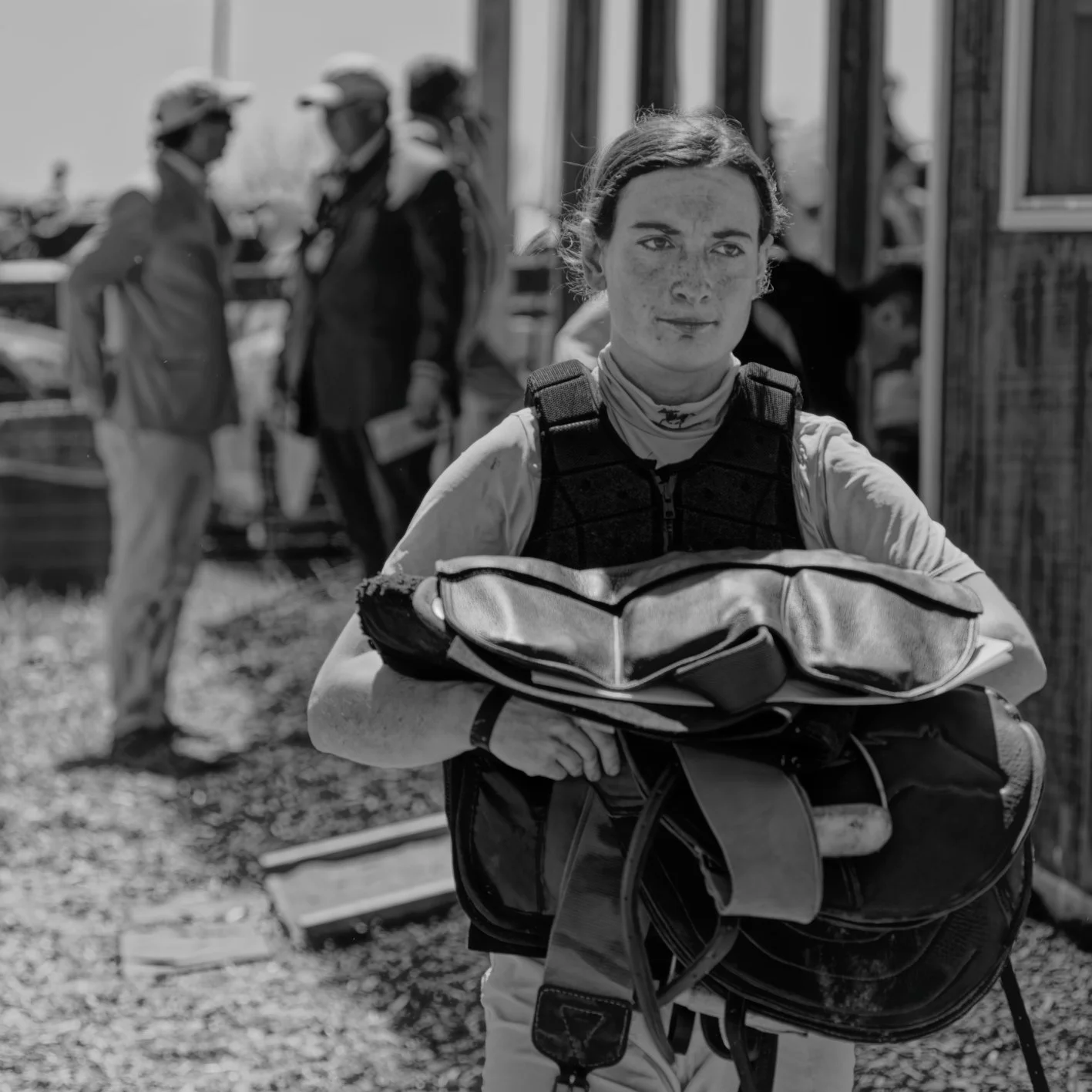 A young woman in race gear carrying a helmet and gear bag, with several people in the background outdoors.