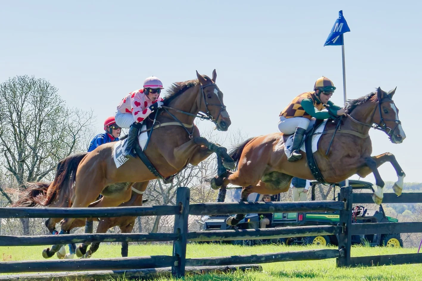 Jockeys in colorful uniforms riding horses over a fence during a horse race on a sunny day.