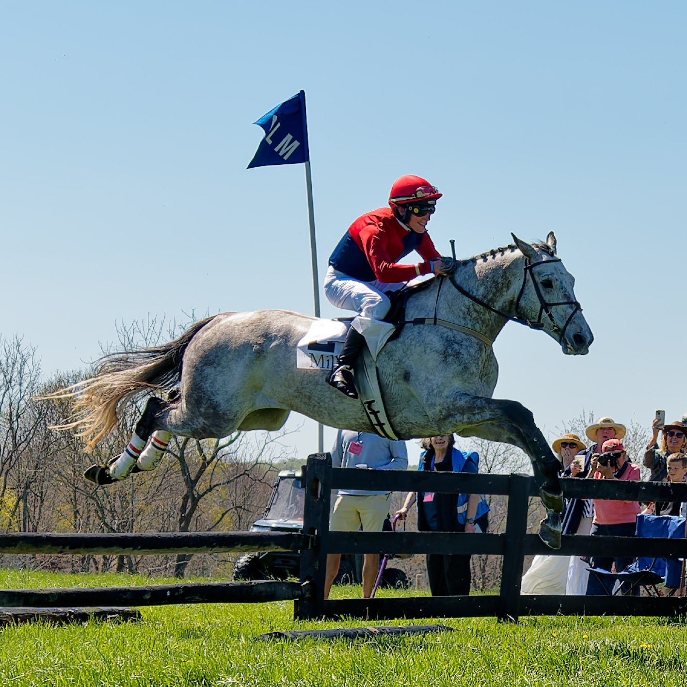 A jockey in red and blue riding attire riding a gray horse over a jump during a cross-country equestrian event, with spectators watching and taking photos in the background under a clear blue sky.