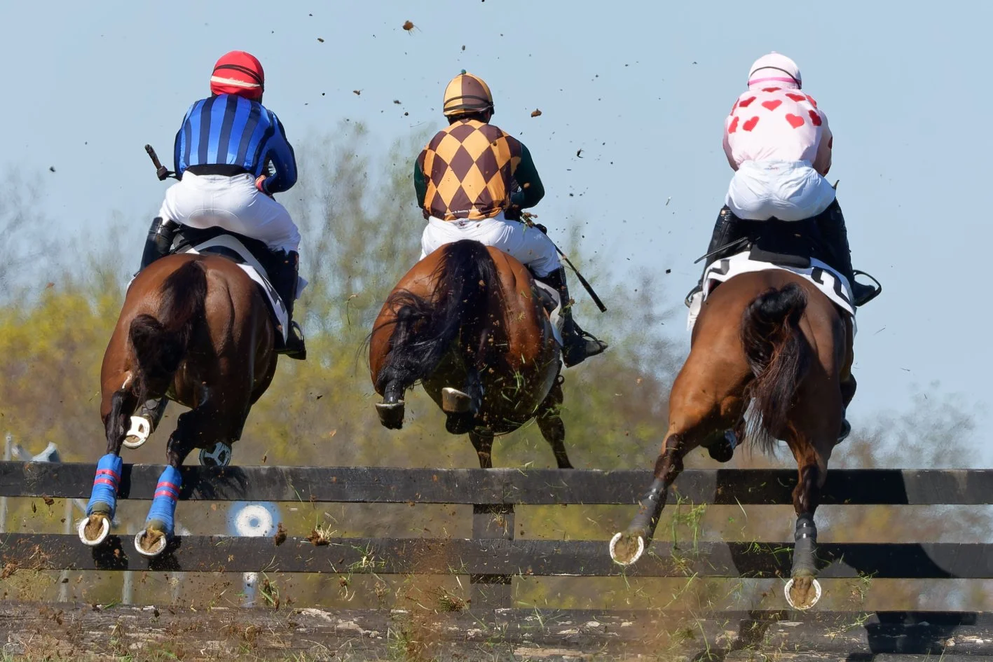 Three jockeys riding horses over a jumping obstacle during a race, with dirt and debris flying in the air.