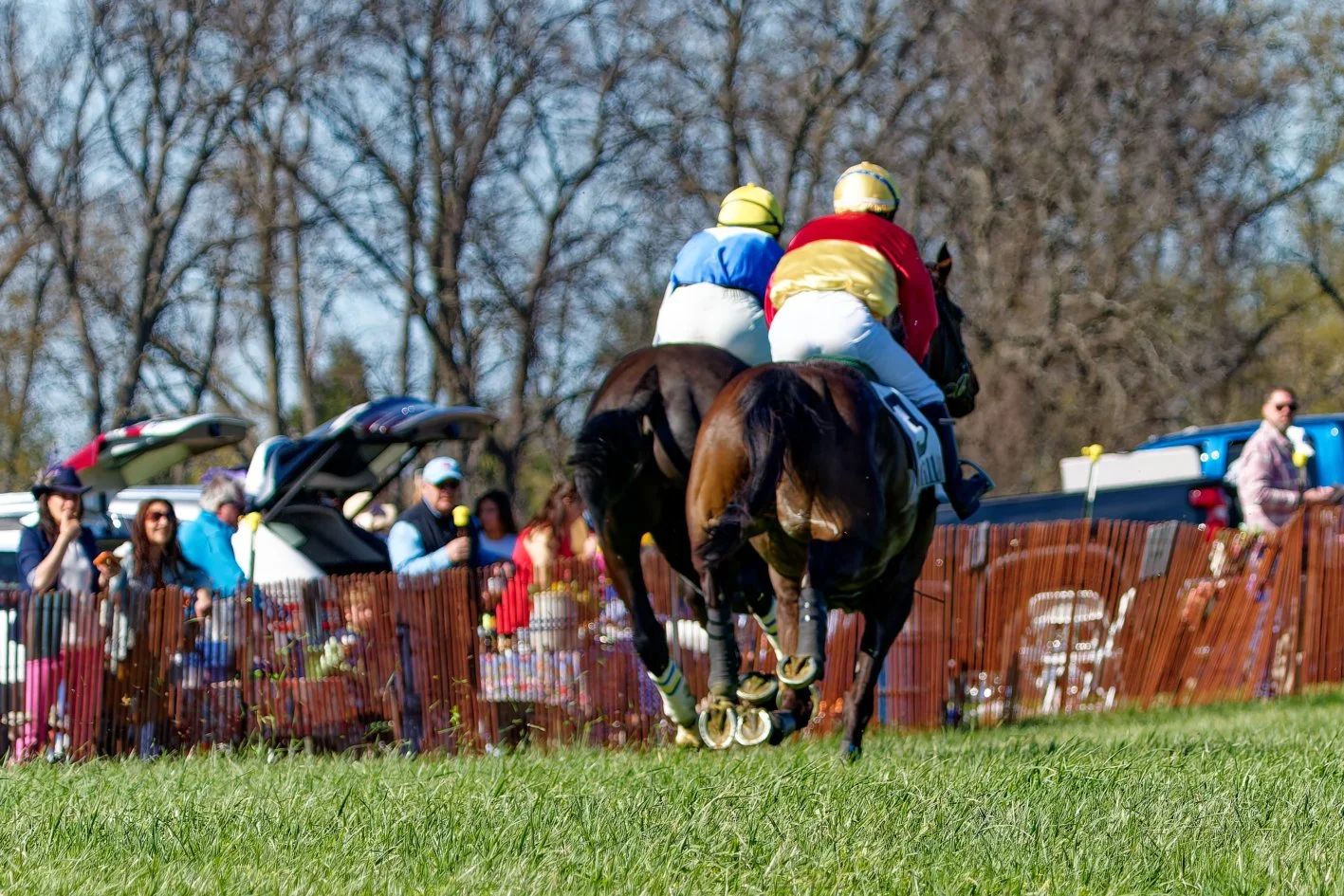Two jockeys riding racehorses on a grassy track, with spectators behind a wooden fence watching and taking pictures.
