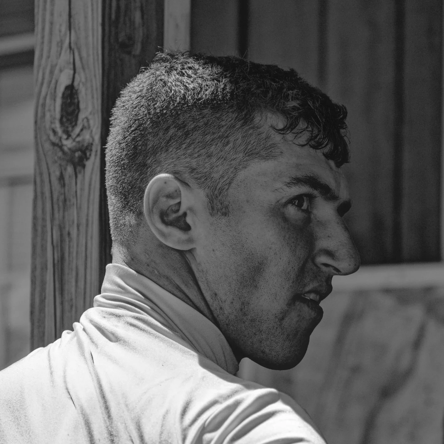 A black and white photo of a man with short hair, wearing a light-colored shirt, looking to the right with an intense expression, standing outdoors near a wooden structure.