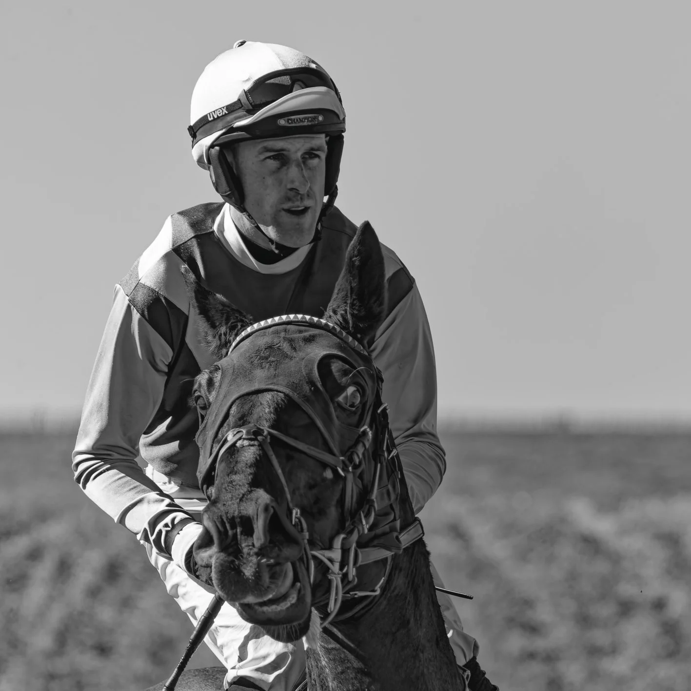 A male jockey wearing a helmet, goggles, and racing silks riding a racehorse during a race or practice session in a flat open field.