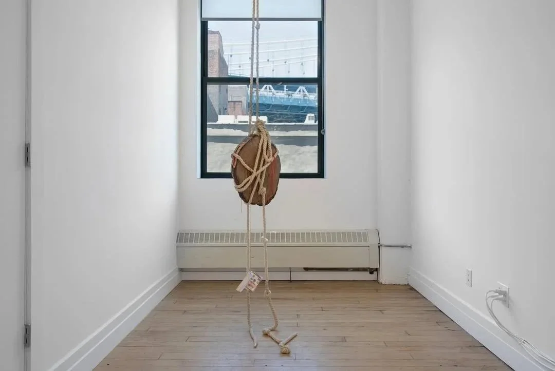 Empty room with white walls, hardwood floor, a window showing a boat outside, and a hanging object made of wood and rope.