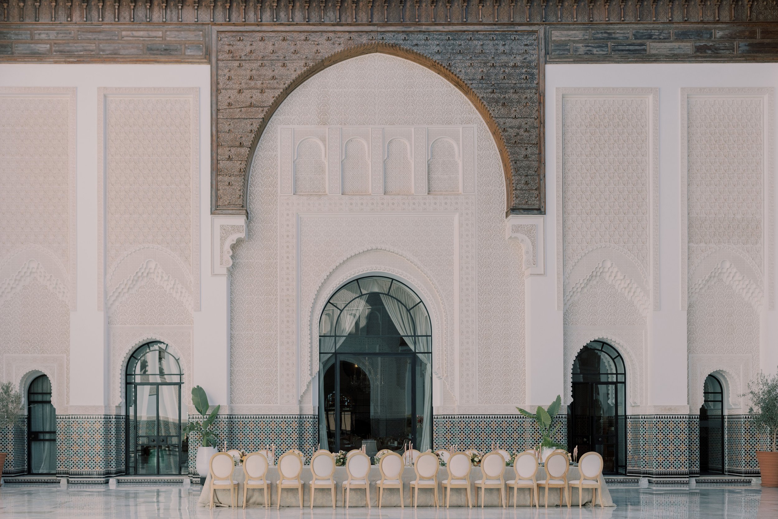 Elegant indoor dining area with a long table and white chairs, ornate white walls with intricate patterns, arched windows, potted plants, and a high decorated ceiling.