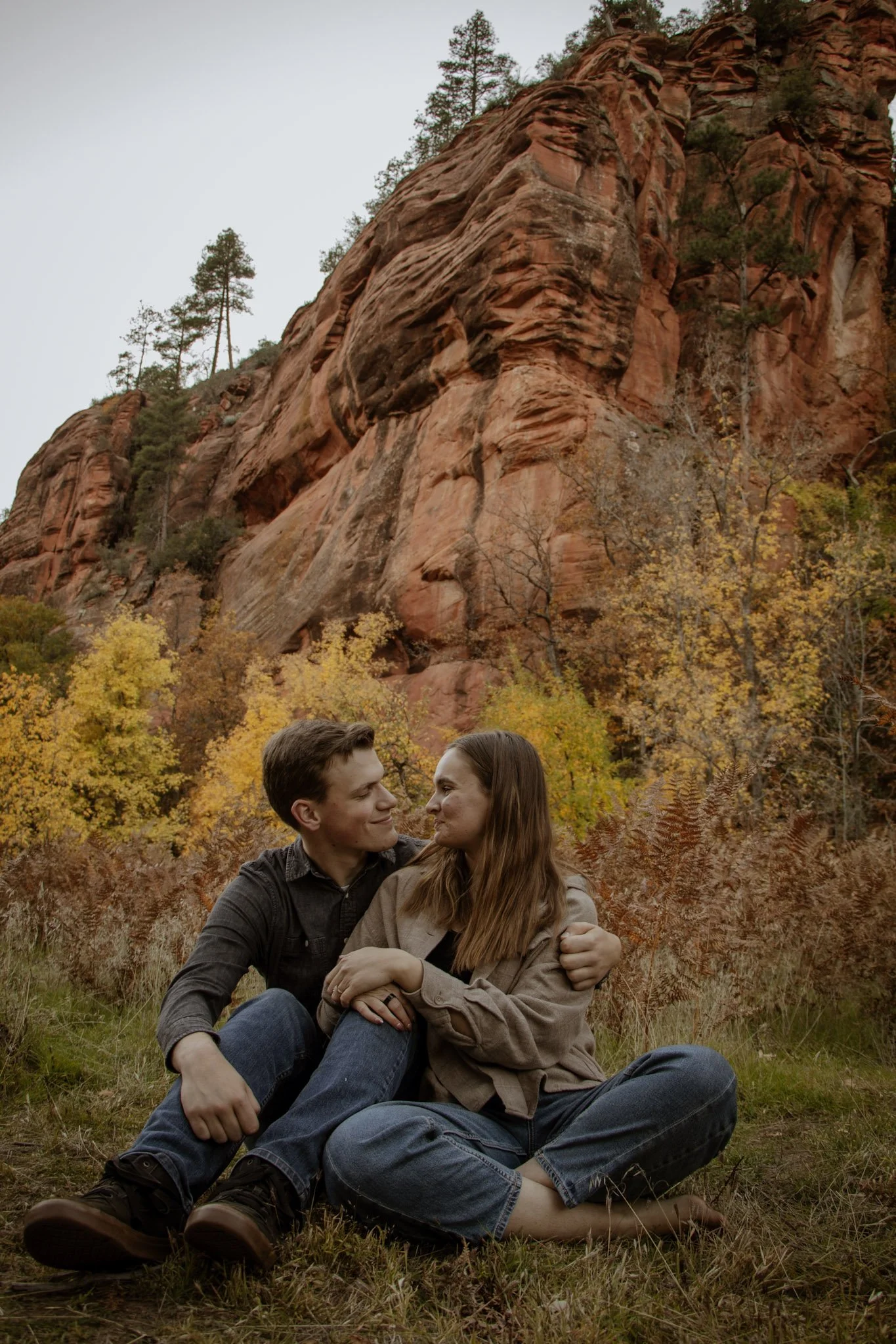 A couple sitting closely together in a grassy area with autumn foliage, trees, and large red rock formations in the background.