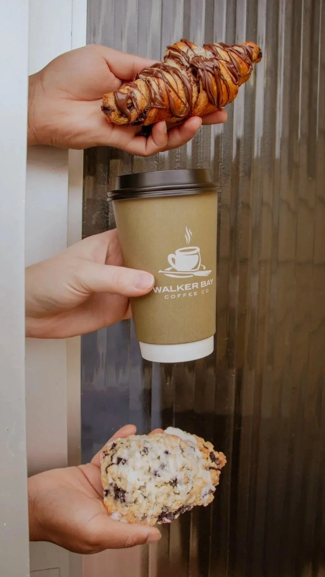 Three hands holding different food items: a croissant with chocolate drizzle at the top, a disposable coffee cup in the middle with 'Walker Bay Coffee Co' logo, and a chocolate chip cookie at the bottom. There is a wooden background behind them.