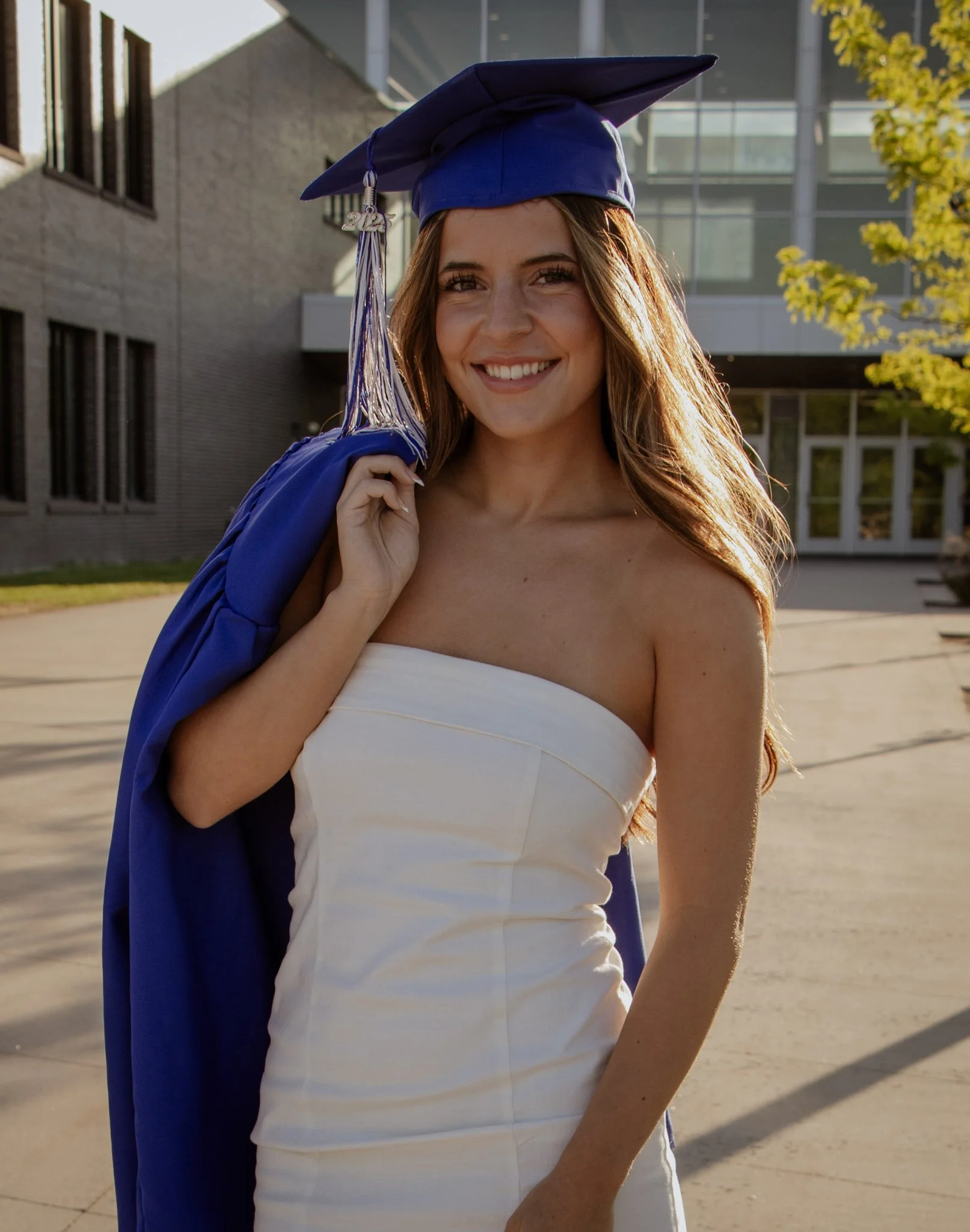 A young woman in a strapless white dress holding a blue graduation gown and cap with a '2023' tassel, smiling outdoors on a sunny day.