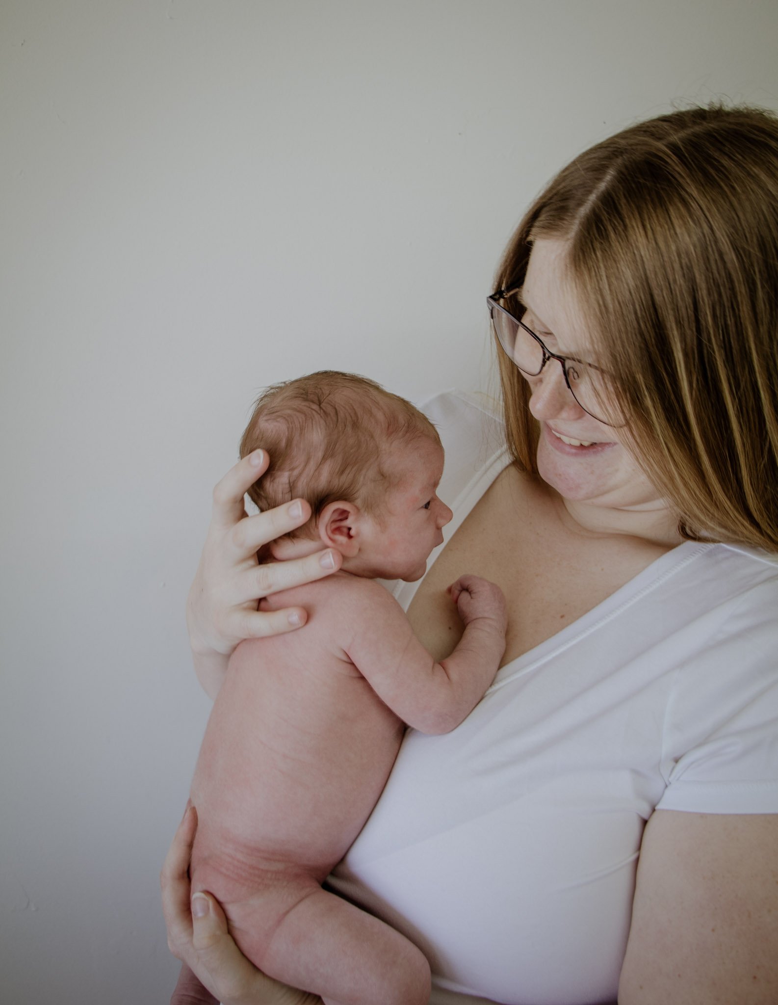 A woman with red hair and glasses holding a newborn baby close to her chest, both with visible skin tones against a plain white background.